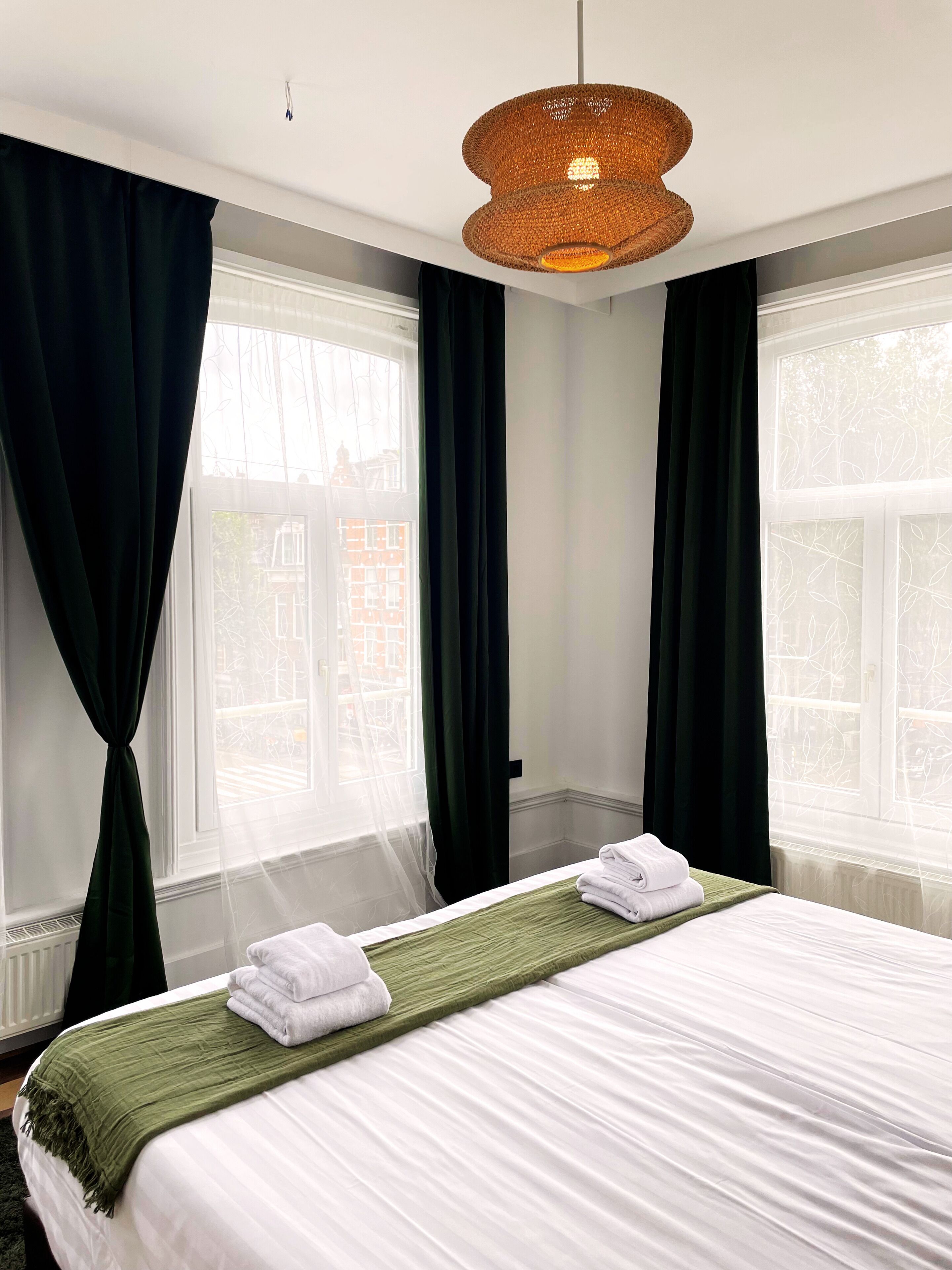 A hotel room with a bed, two windows with curtains, a hanging light fixture, and a view of buildings outside.