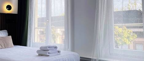 Un lit bien fait, avec du linge de lit blanc et une serviette pliée, un applique ronde au mur et une fenêtre donnant sur des immeubles et des arbres.