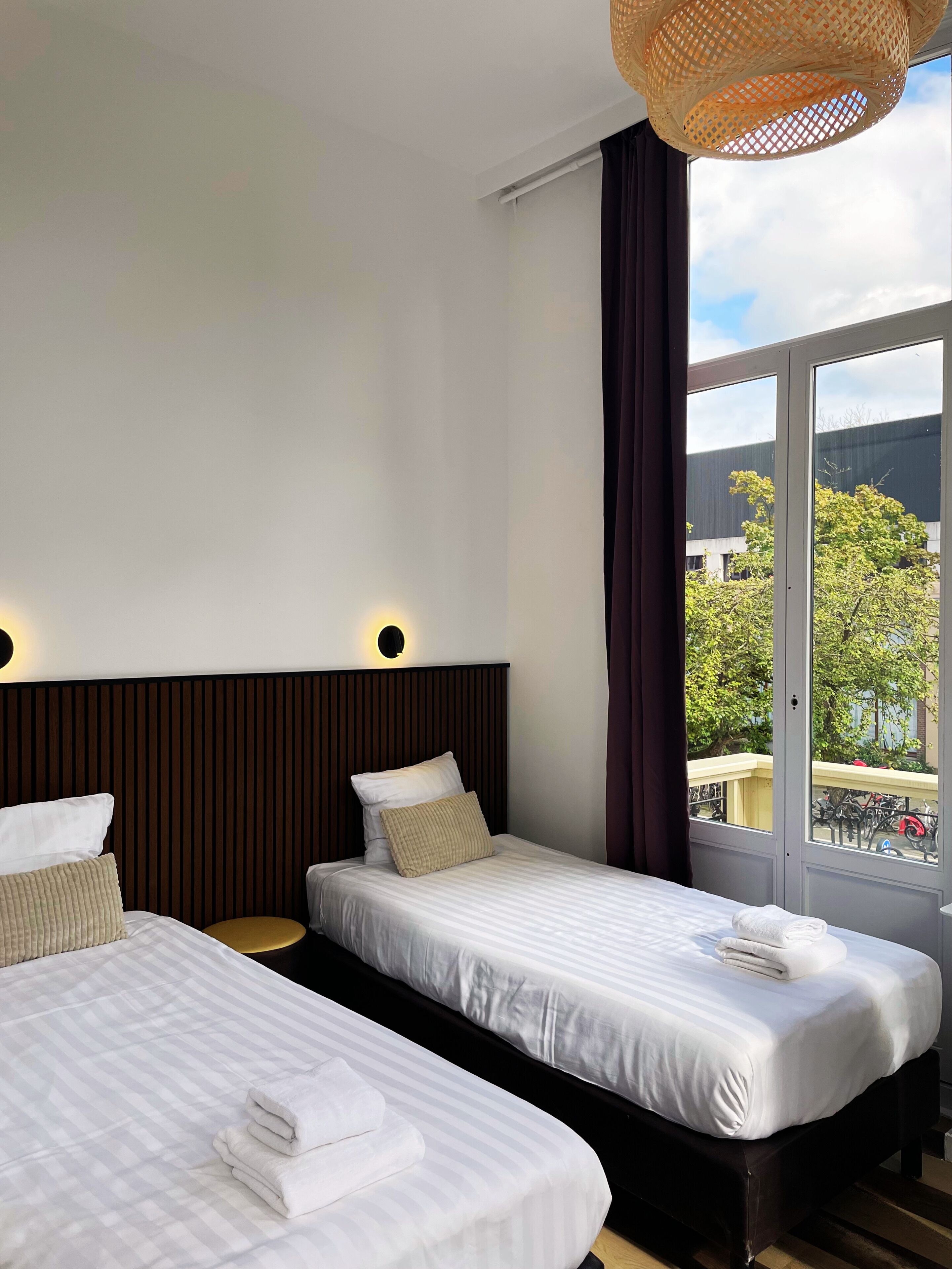 A hotel room with two beds, a large window with a view, and a woven light fixture.