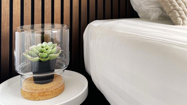 A neatly made bed with white bedding and a beige pillow, a small round table with a glass terrarium containing a succulent, and a wooden slat wall.