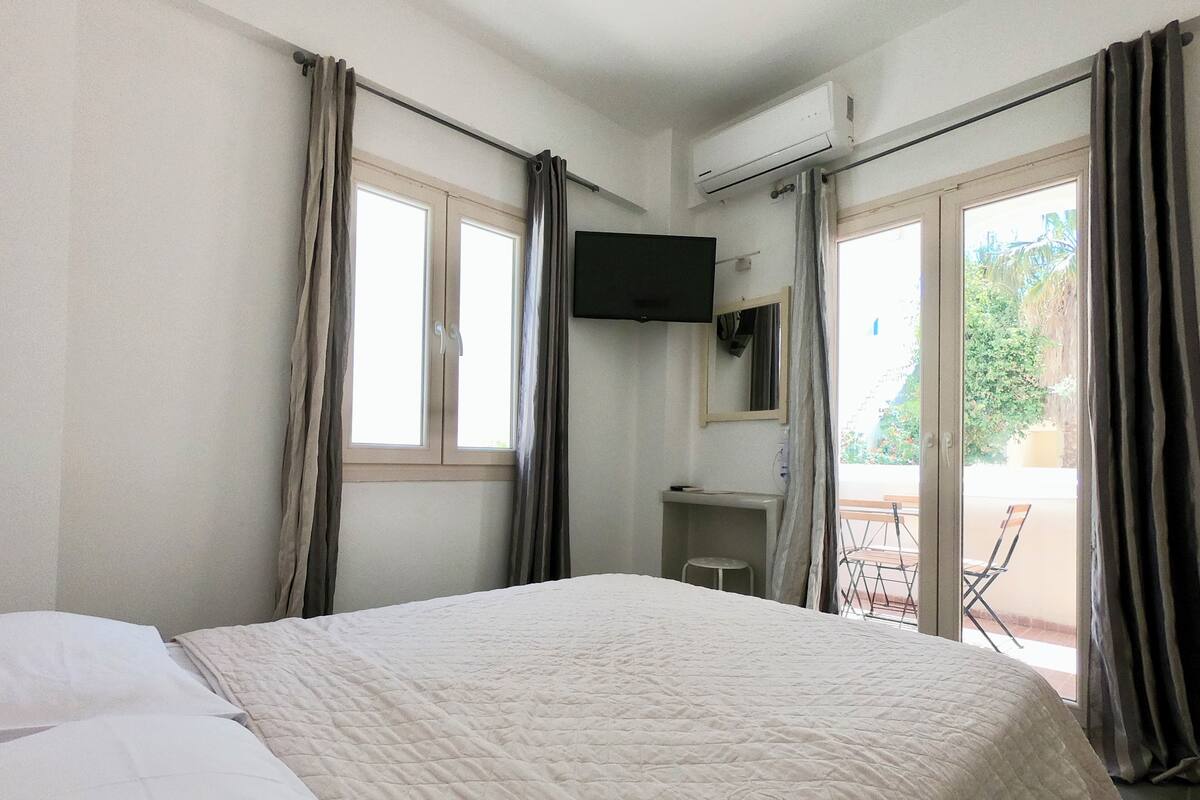 Habitación de hotel con cama, televisor montado en la pared, escritorio pequeño con silla y balcón con mesa y sillas.