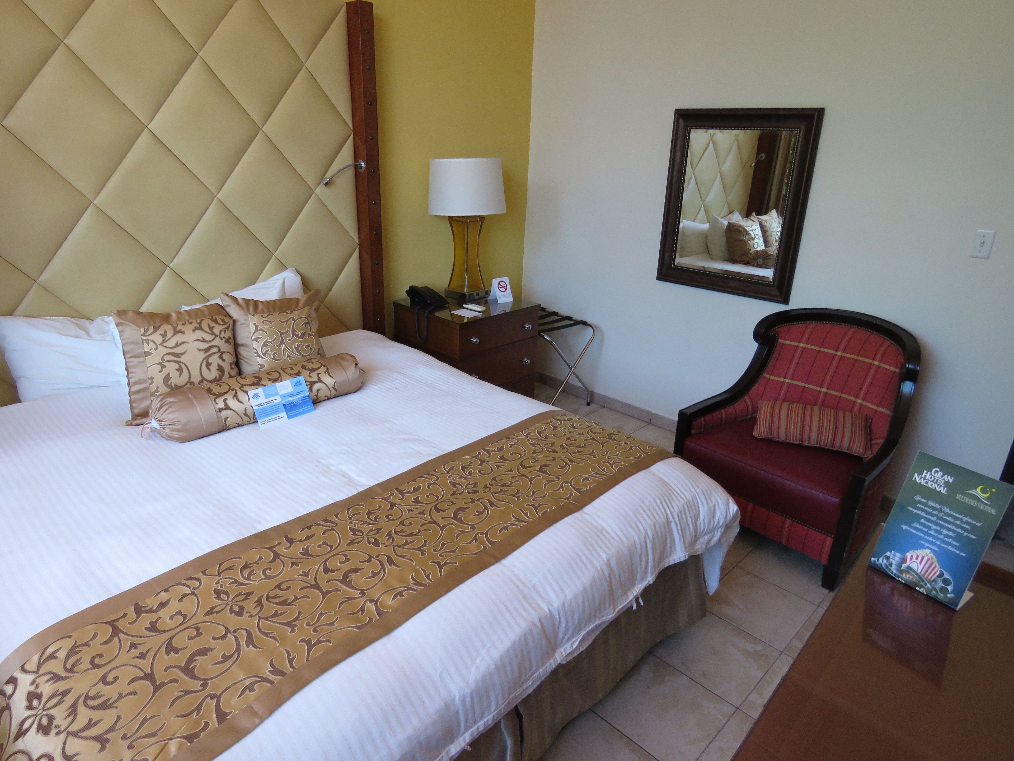 Habitación de hotel con una cama grande, un sillón rojo, una mesita de noche, una lámpara y un espejo en la pared.
