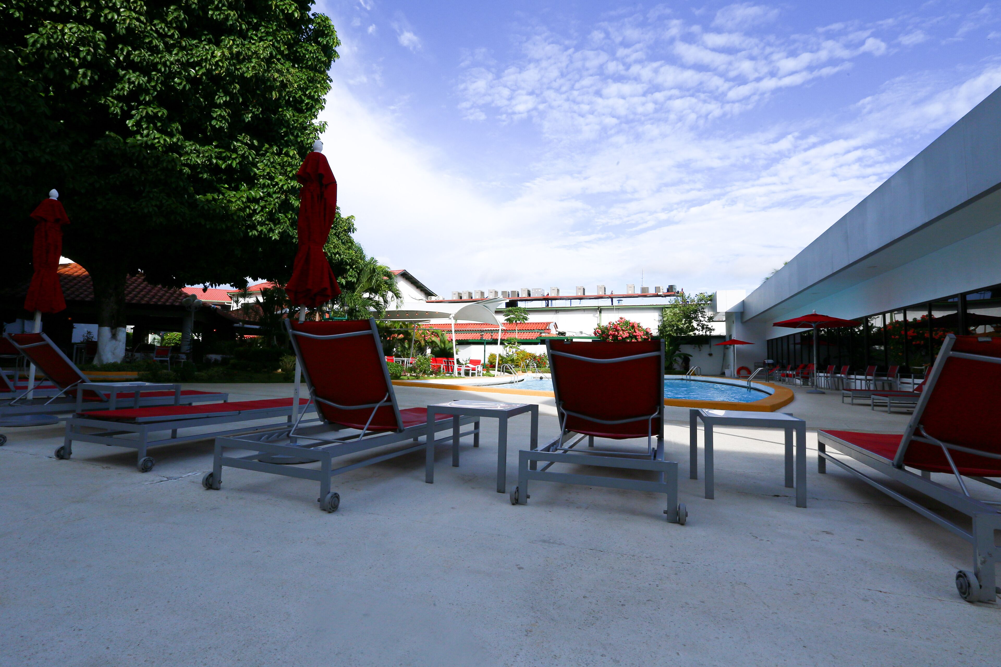 Zona de piscina con tumbonas y sombrillas, cielo despejado y un edificio con tejado rojo al fondo.