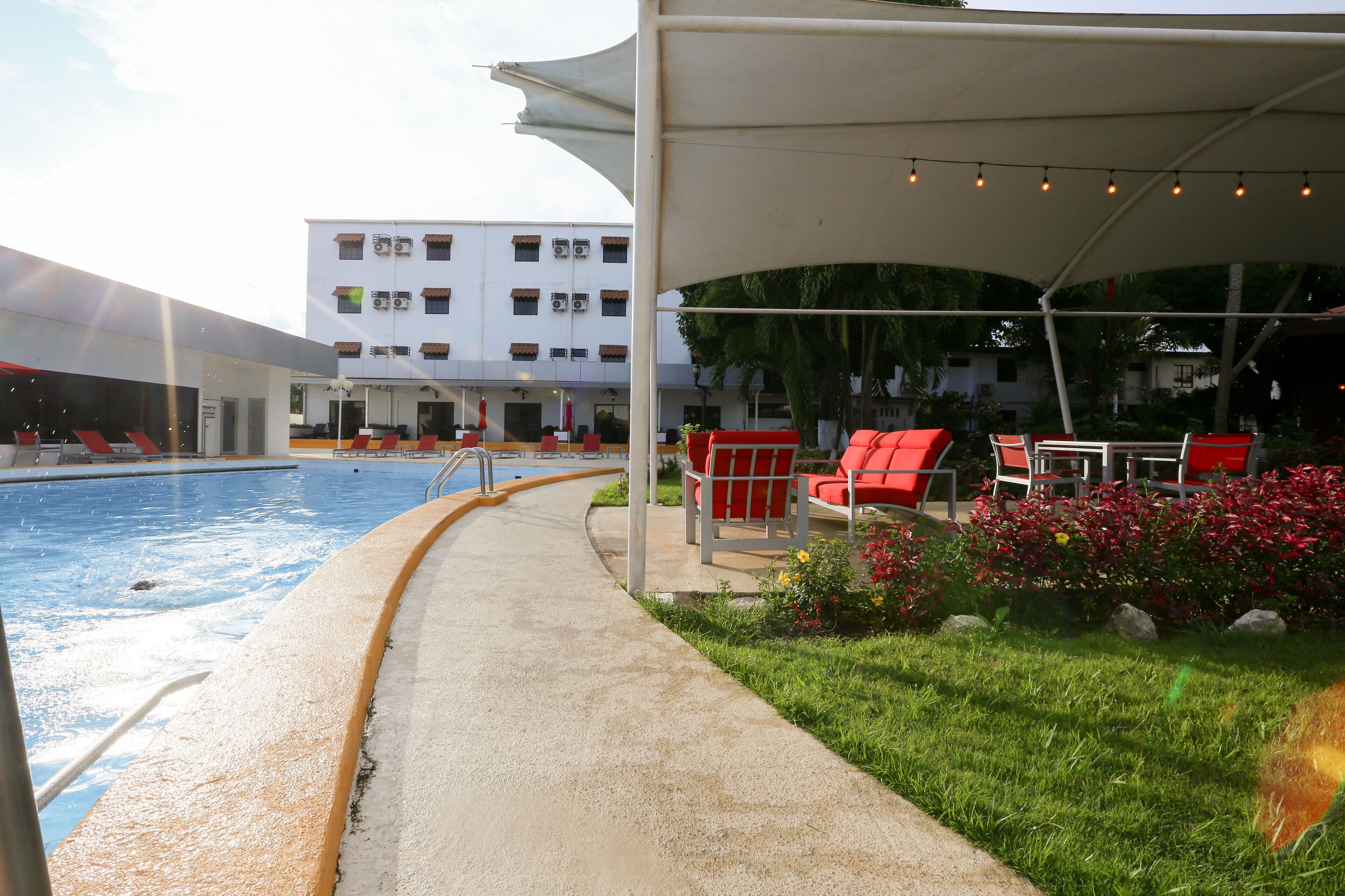 A poolside area with a concrete walkway, red chairs, and a covered seating area with string lights.