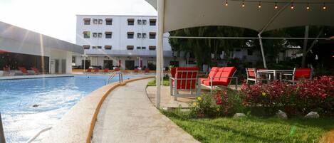 A poolside area with a concrete walkway, red chairs, and a covered seating area with string lights.