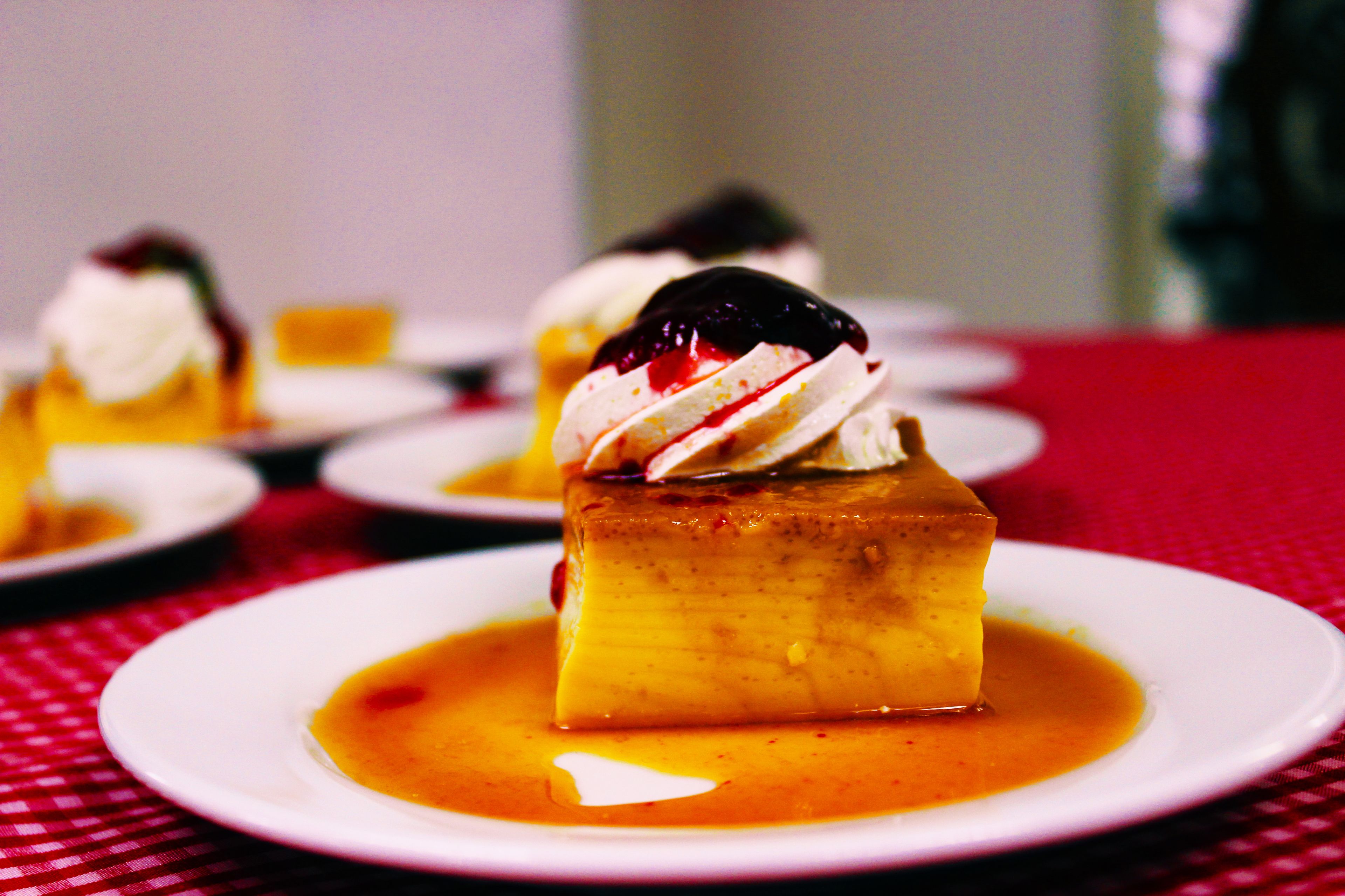 Un postre con crema batida y una cereza encima, servido en un plato blanco con salsa de caramelo, sobre un mantel a cuadros rojos y blancos.