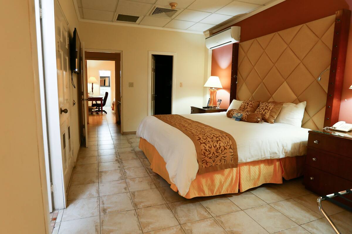 Una habitación de hotel con una cama grande, una mesita de noche con lámpara, un aparador y una puerta que da a un pasillo.