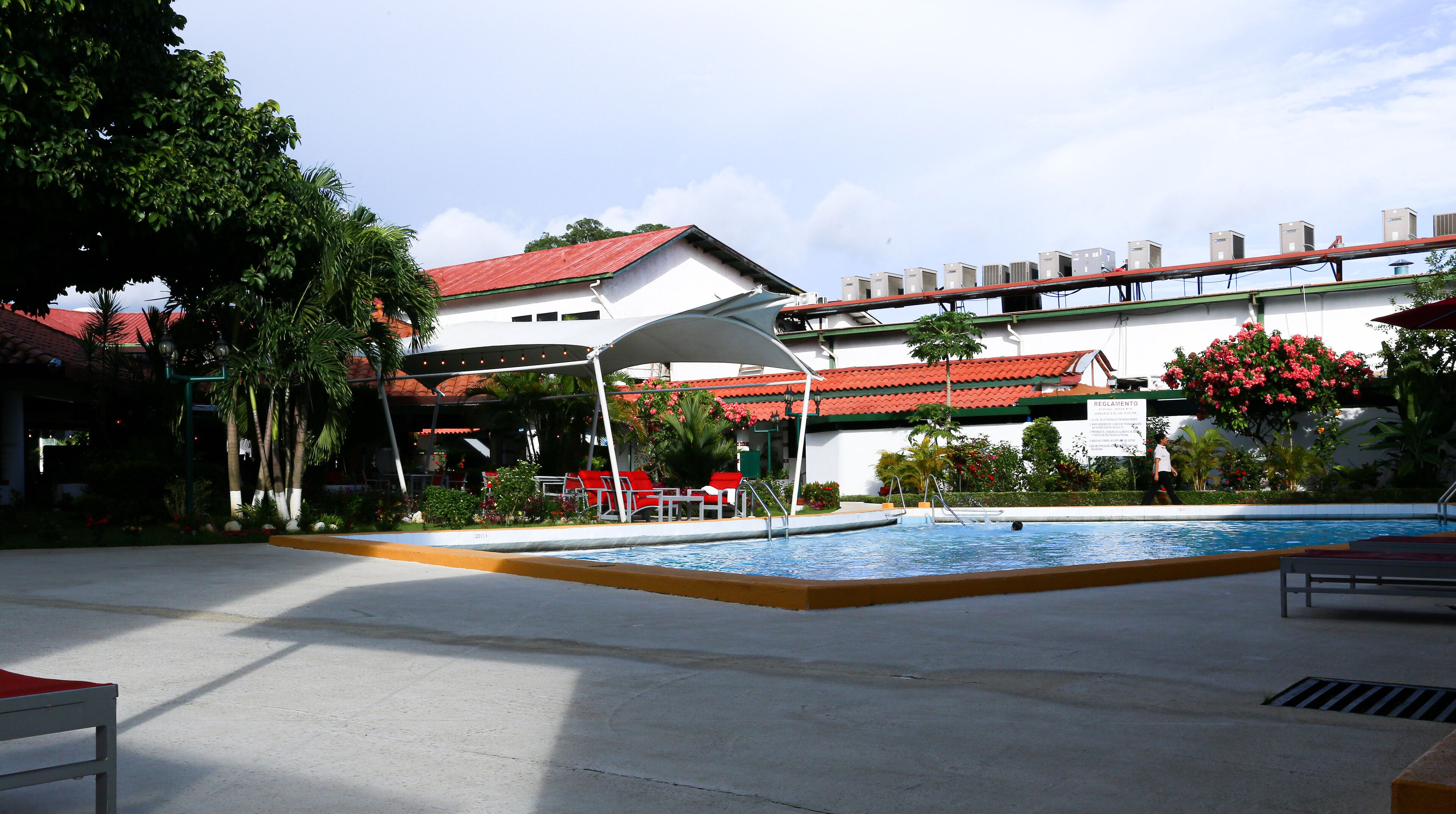 Una piscina con sedie rosse e una zona salotto coperta, immersa nel verde e con un edificio dal tetto rosso.