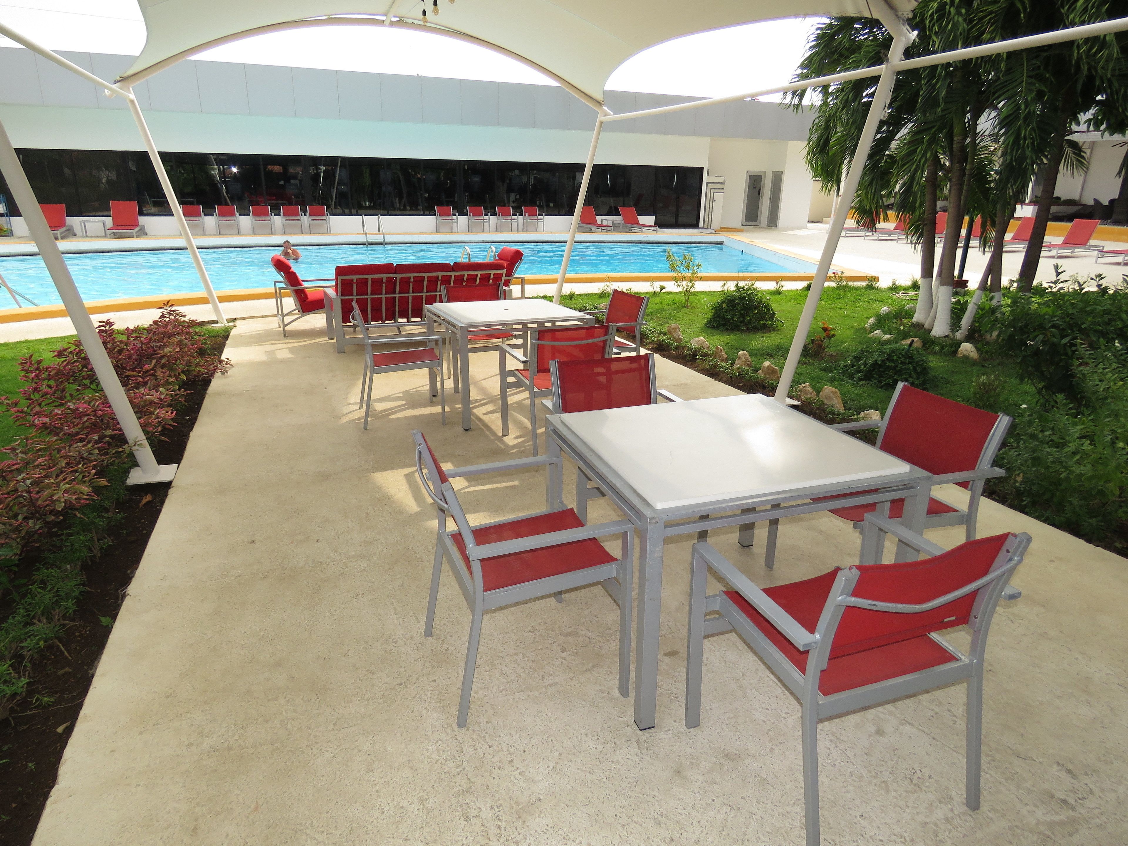 Uma área de piscina ao ar livre com espaço de estar coberto, cadeiras vermelhas e uma mesa branca.