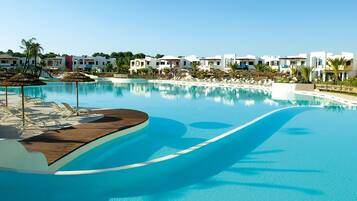 A large, clear swimming pool with lounge chairs and umbrellas, surrounded by modern buildings with balconies.