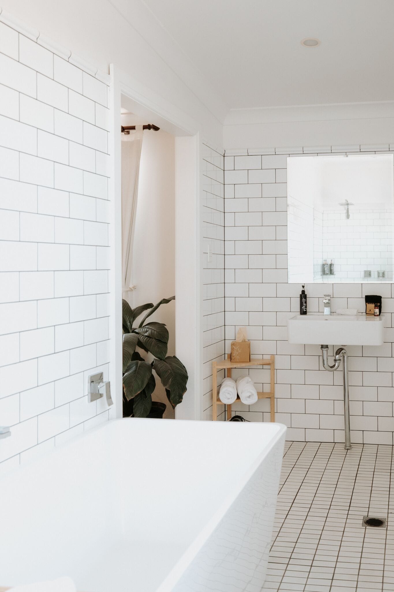 A modern bathroom with white subway tiles, a white bathtub, a wooden shelf with towels, and a mirror above the sink.