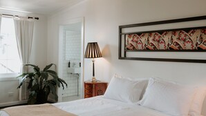 Een netjes opgemaakt bed met witte lakens, een bedlampje en een lijst met een kunstwerk erboven.