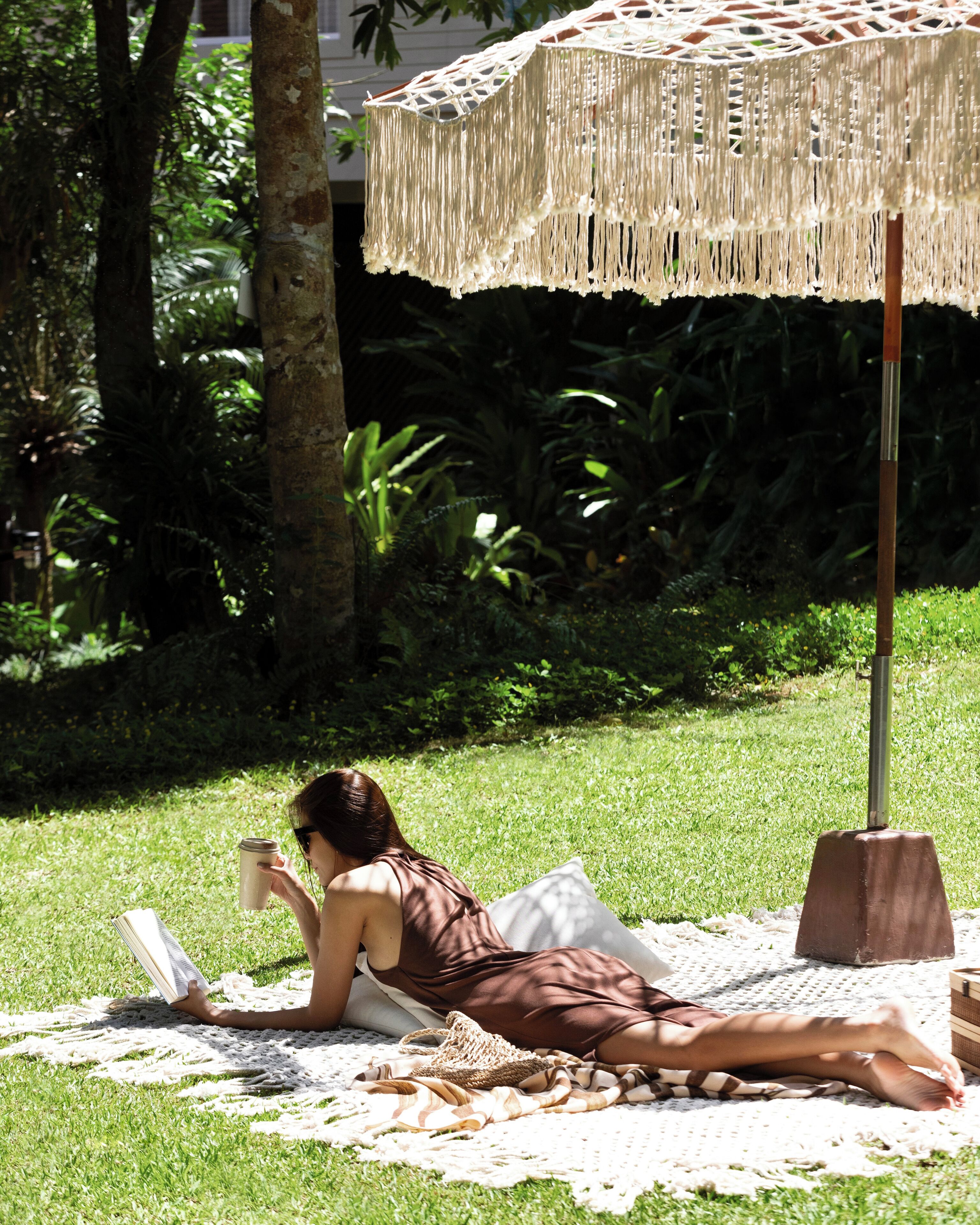 A woman reading a book on a blanket under a macrame umbrella in a grassy area with trees.