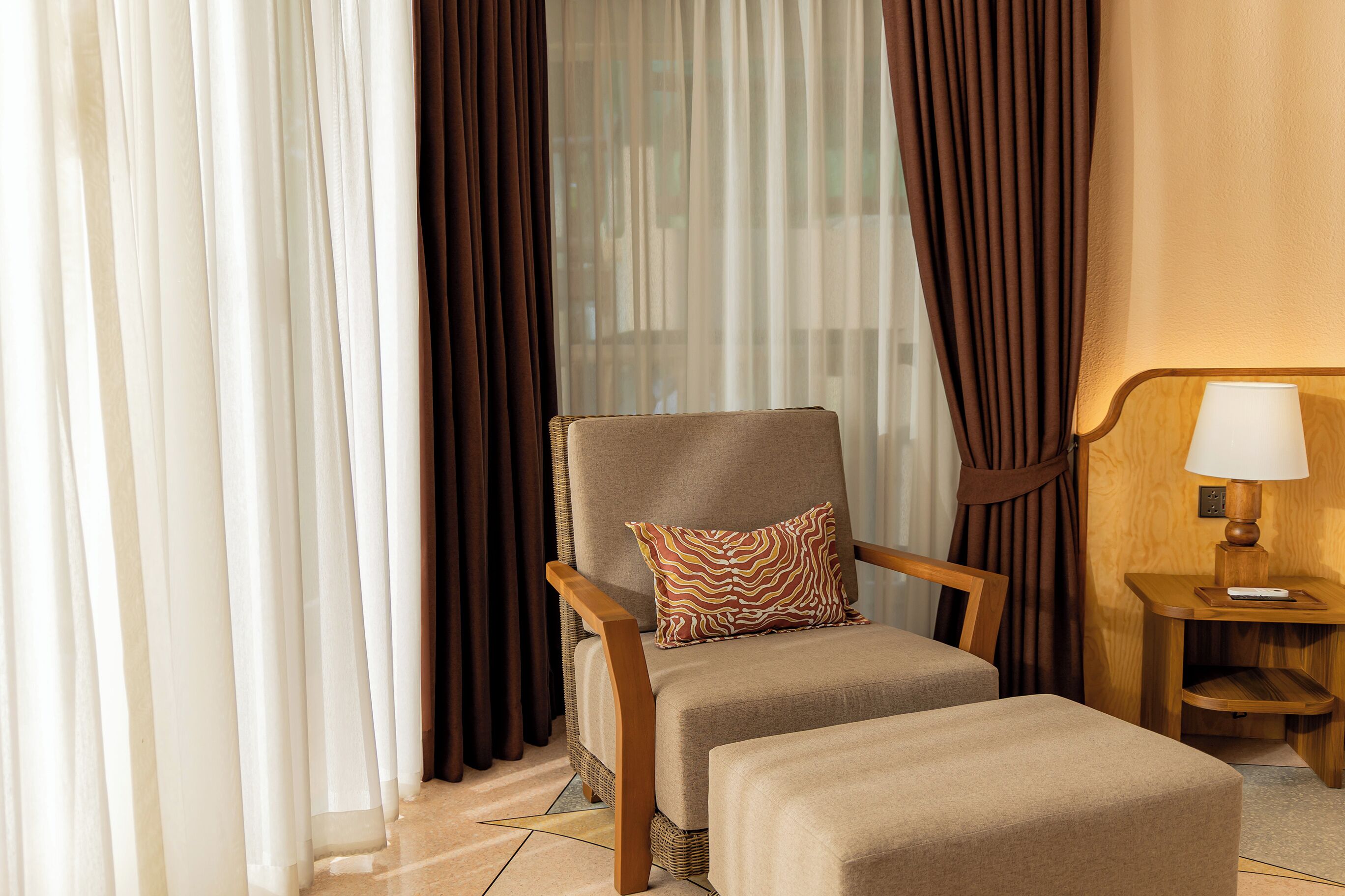 Habitación de hotel con un sillón de madera, un reposapiés a juego, una lámpara sobre una mesita de madera y cortinas de encaje.