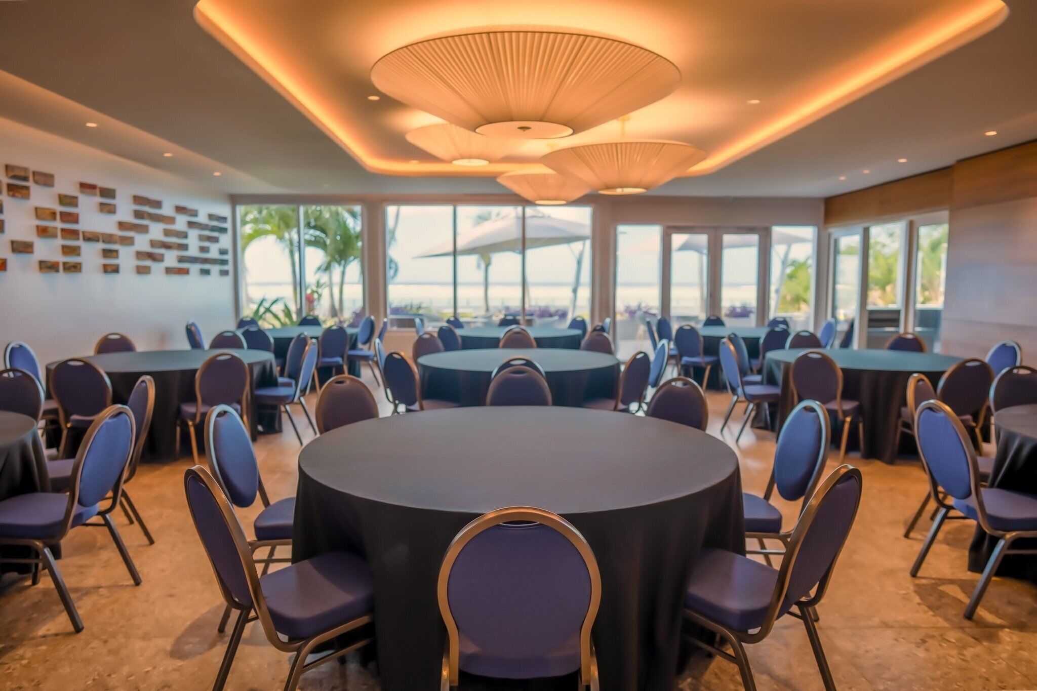 A conference room with multiple round tables and chairs, large windows, and a modern ceiling design.