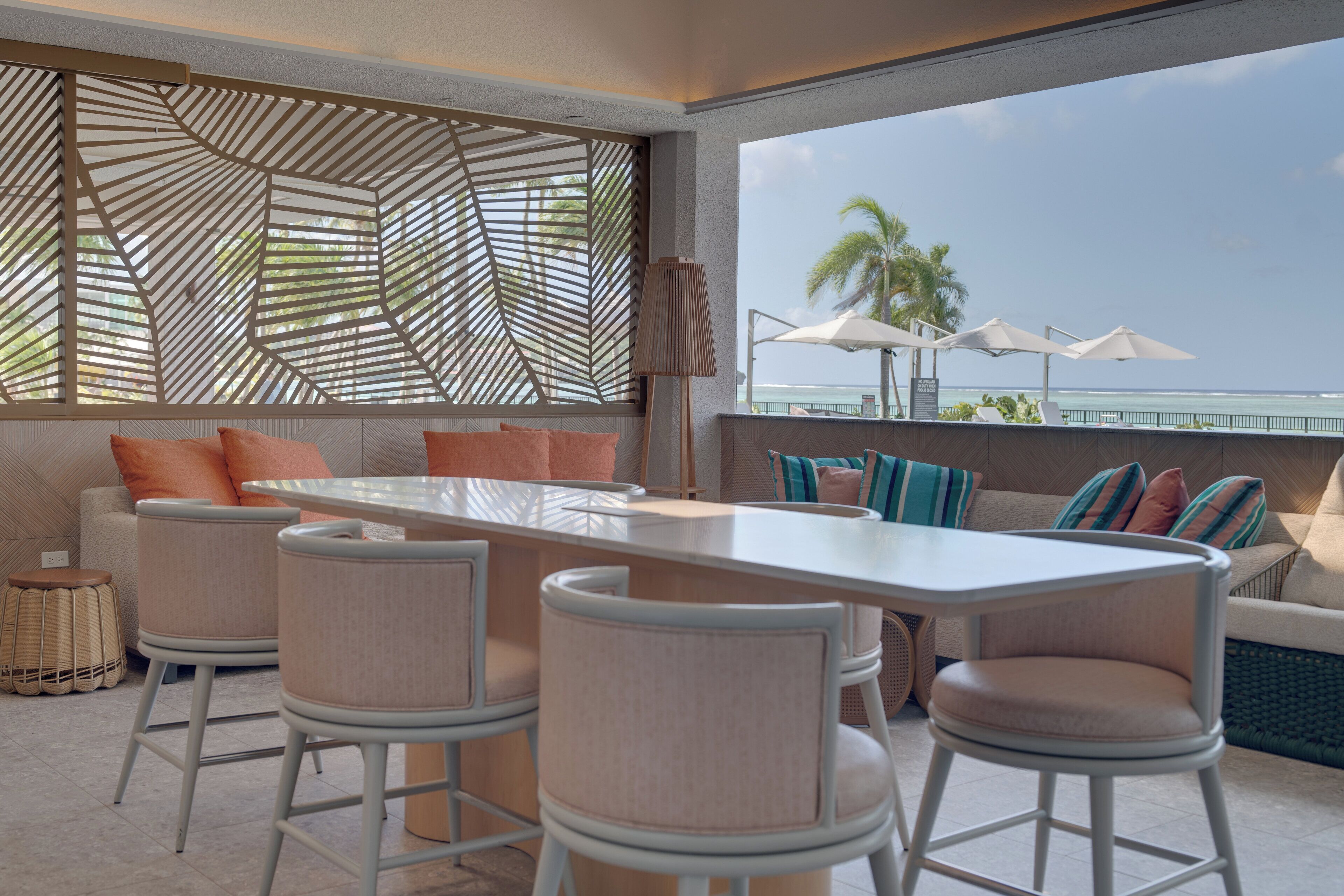 Un área de estar al aire libre y de estilo moderno con una mesa larga y taburetes, con vista a una playa.