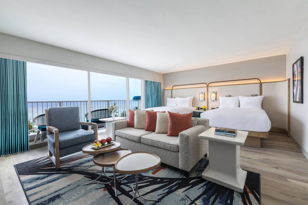 A modern hotel room with a bed, a sofa, a chair, a small table, and a view of the ocean.