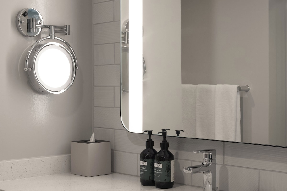 A modern bathroom with a white sink, a round mirror, a towel rack, and two bottles of body wash.