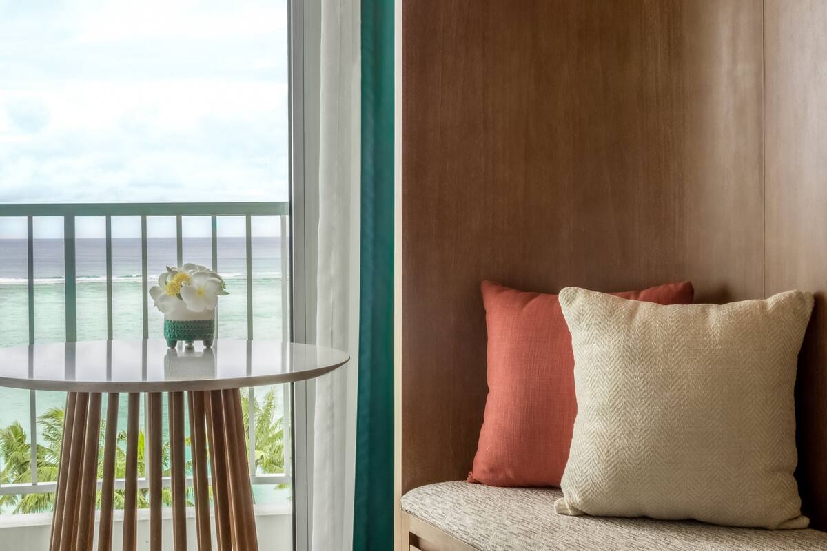 A balcony with a view of the ocean, a round table with a flower, and a cushioned seating area with pillows.