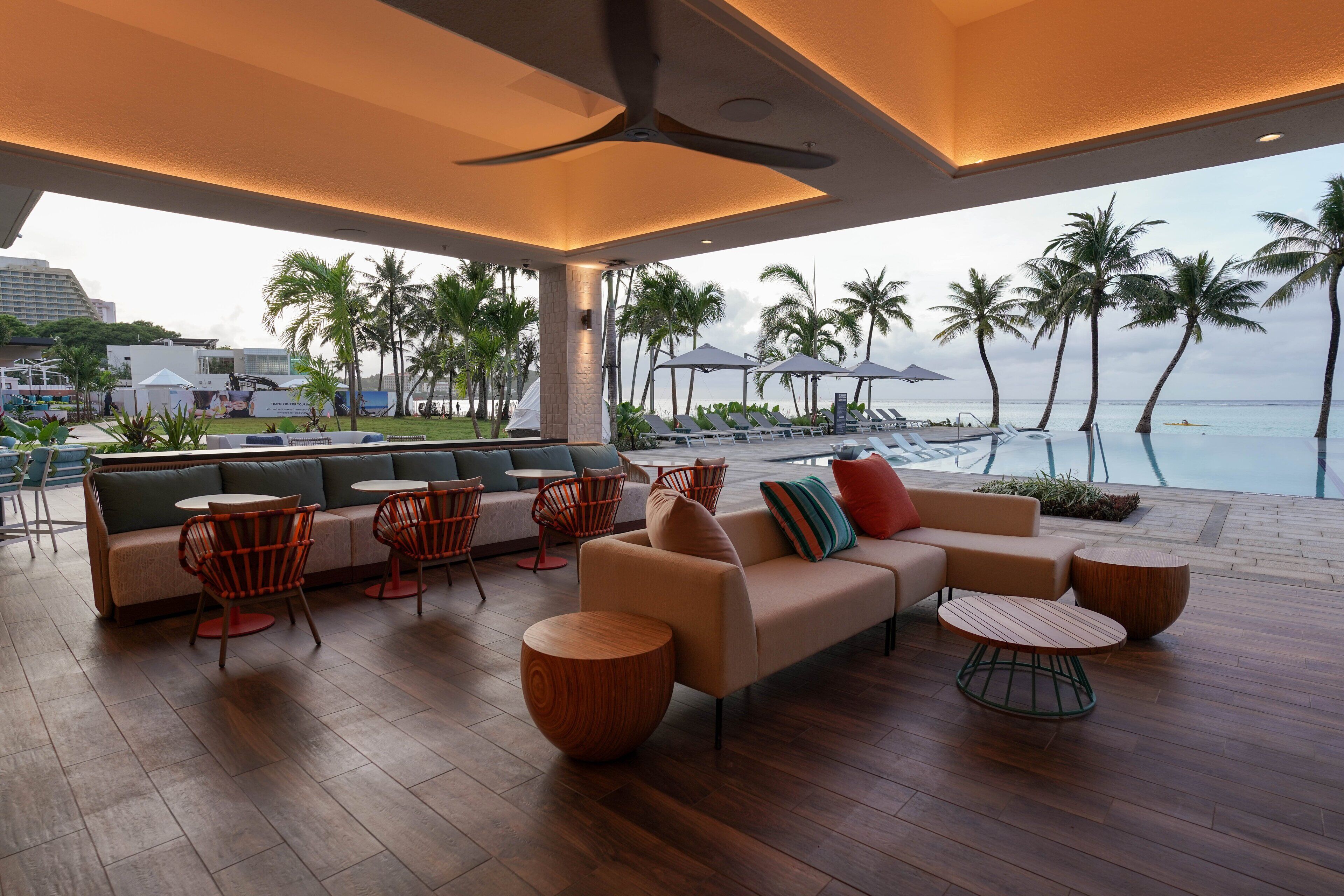 Un espace de détente extérieur moderne, doté d’un canapé, de fauteuils et de tables rondes, offrant une vue sur une plage bordée de palmiers et sur l’océan.