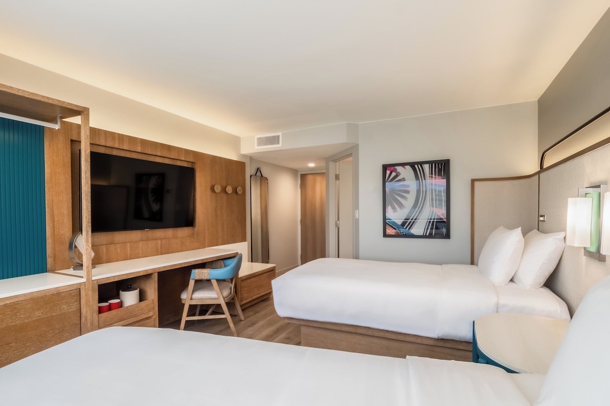 A hotel room with two beds, a desk, a chair, a TV, and a bathroom door.