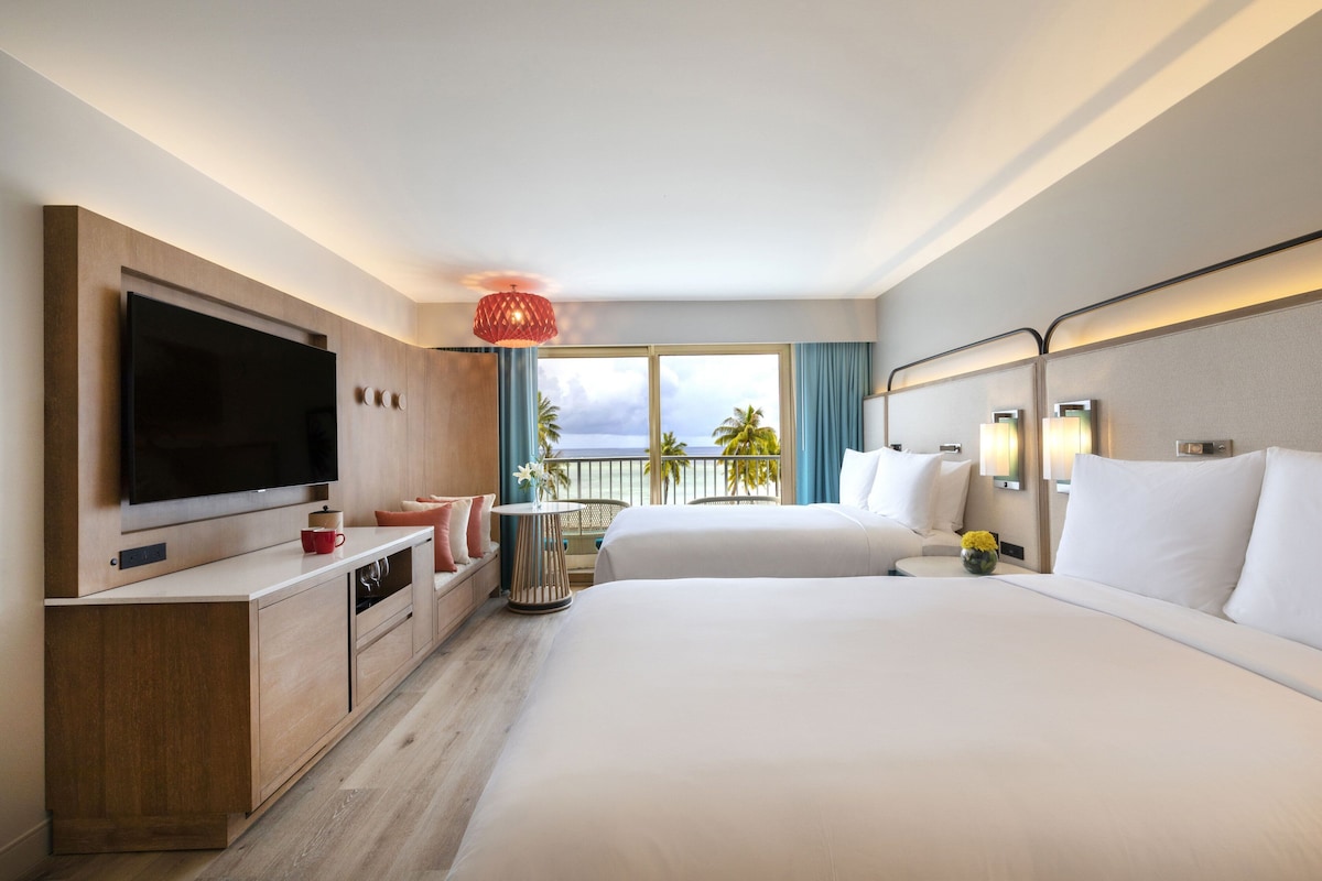 A hotel room with a large bed, a TV, a desk, and a balcony with a view of the ocean.