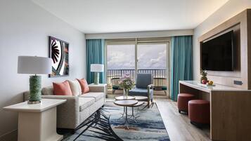 Una habitación de hotel moderna con sofá, dos mesitas auxiliares, una mesa de centro, televisor y balcón con cortinas.