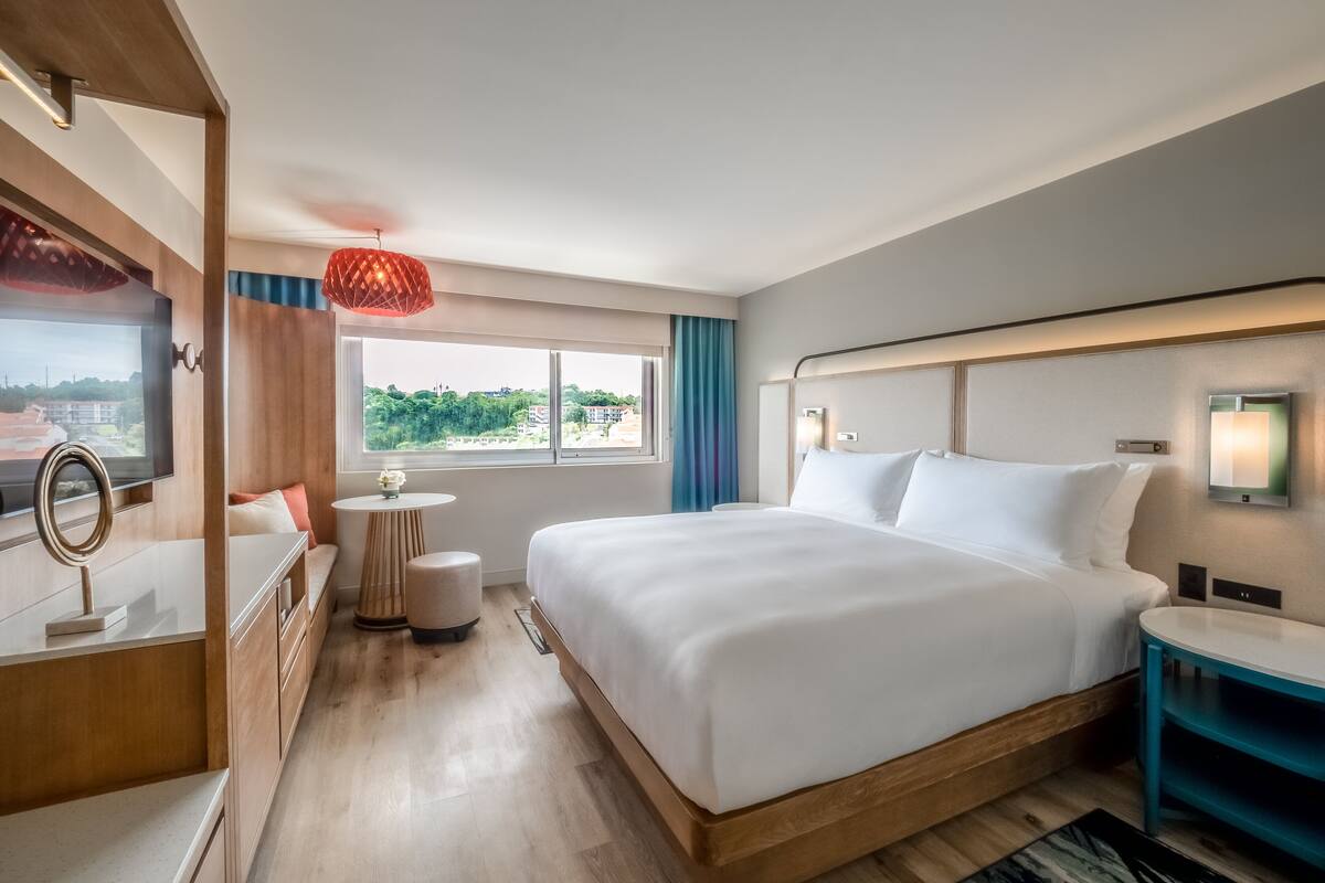 A hotel room with a large bed, a bedside table, a small round table with a lamp, a TV, and a view of greenery outside.