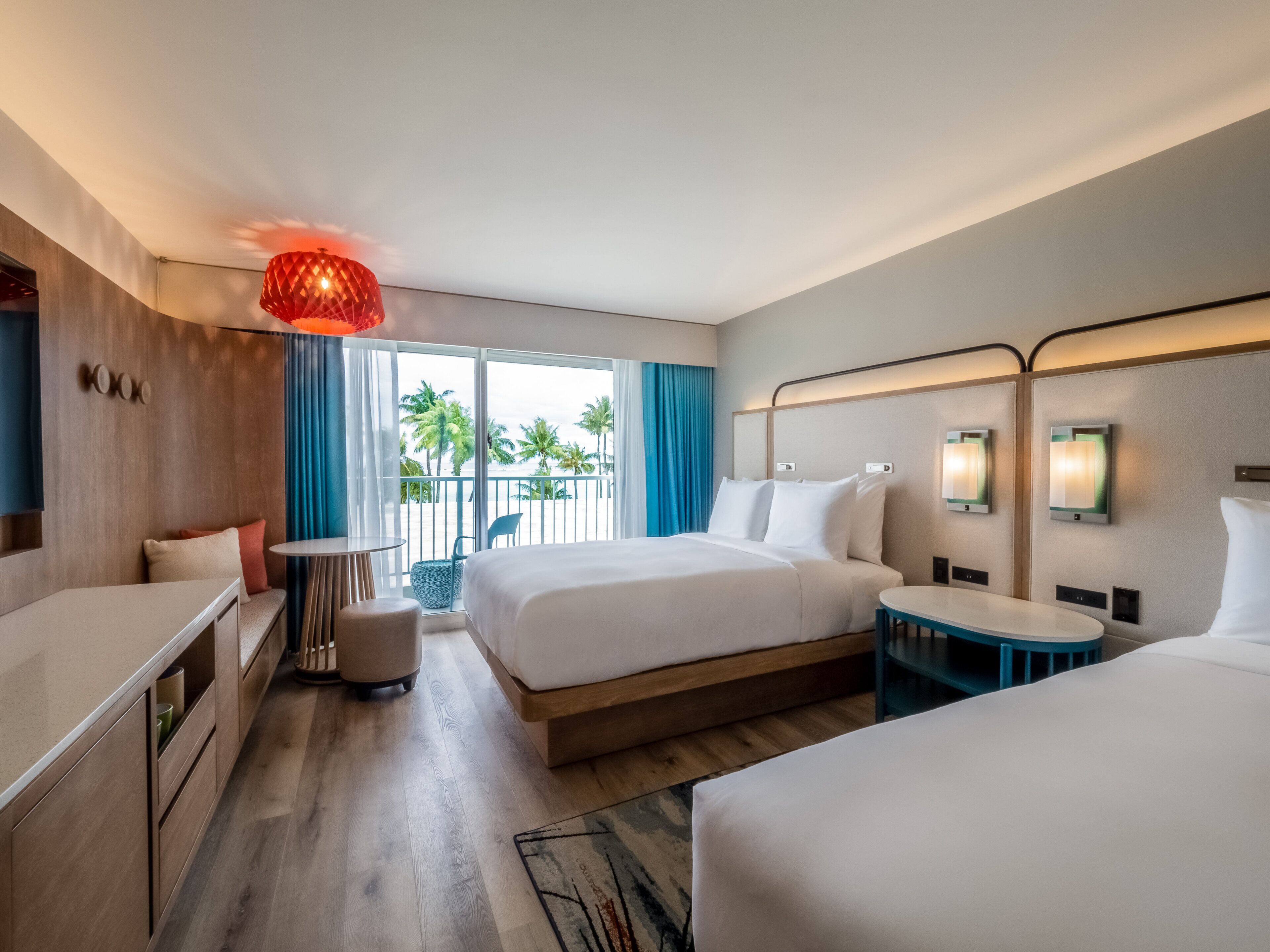 A hotel room with a large bed, a smaller bed, a desk, and a chair. There is a balcony with a view of palm trees and the ocean.