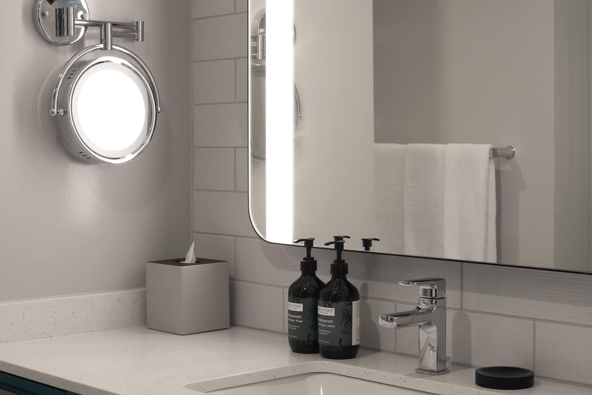 A modern bathroom with a white sink, a large mirror, and a toilet with a round mirror above it. There are two bottles of shampoo and a tissue box on the counter.