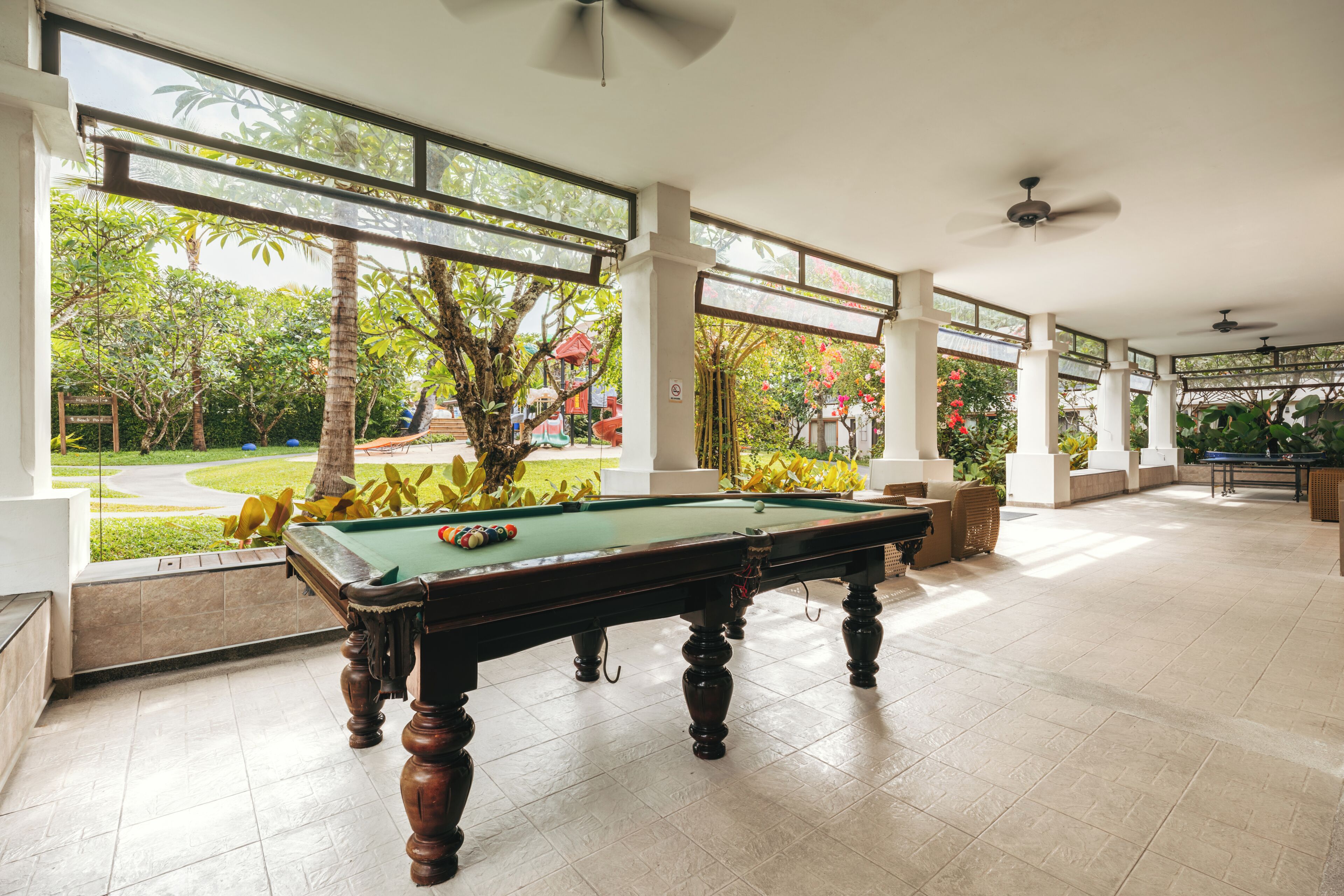 Une table de billard dans une pièce spacieuse, dotée de grandes fenêtres, de ventilateurs de plafond et offrant une vue sur un jardin aménagé.