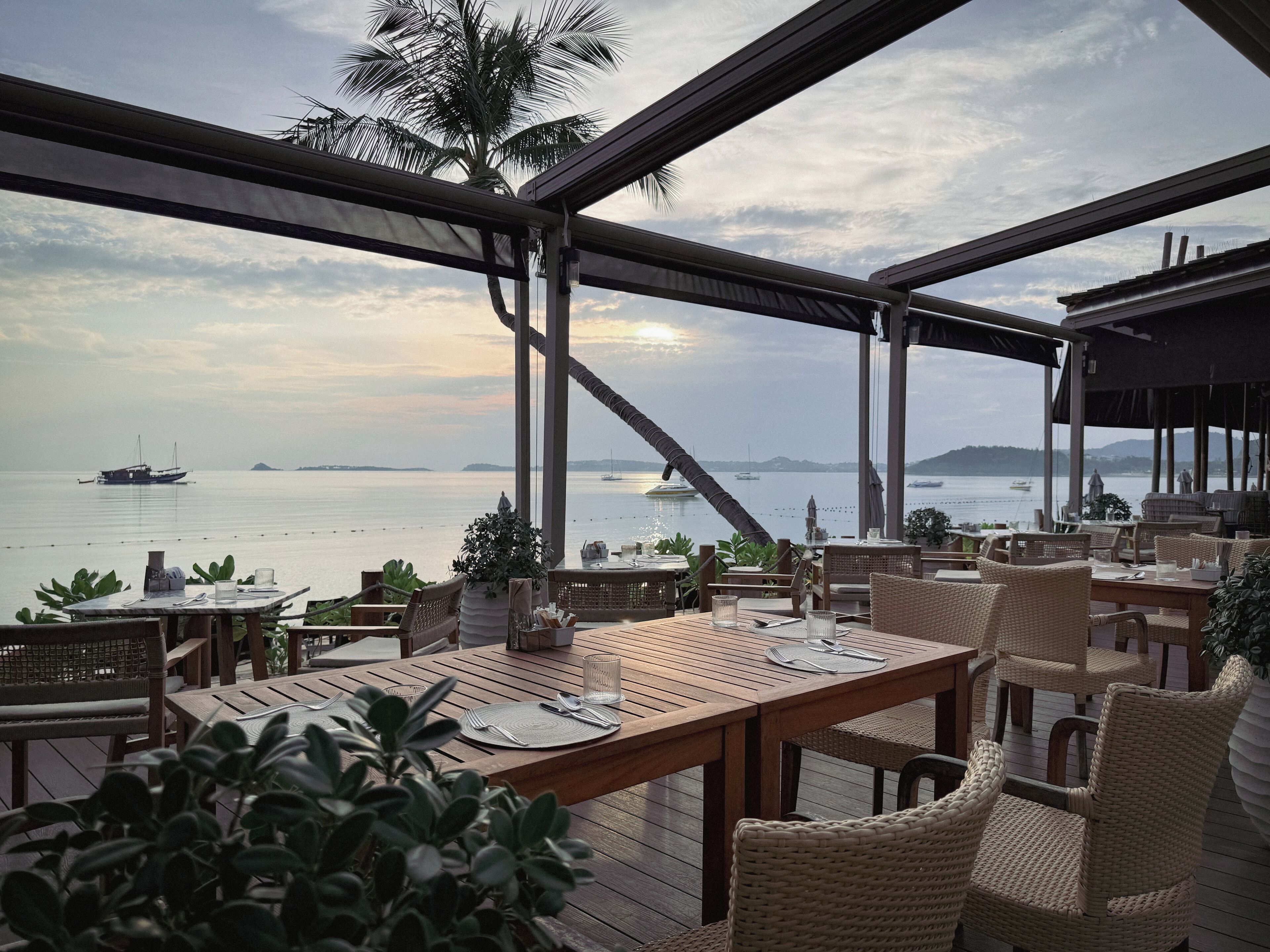 An outdoor dining area with wooden tables and wicker chairs, overlooking a calm sea with boats and distant islands.