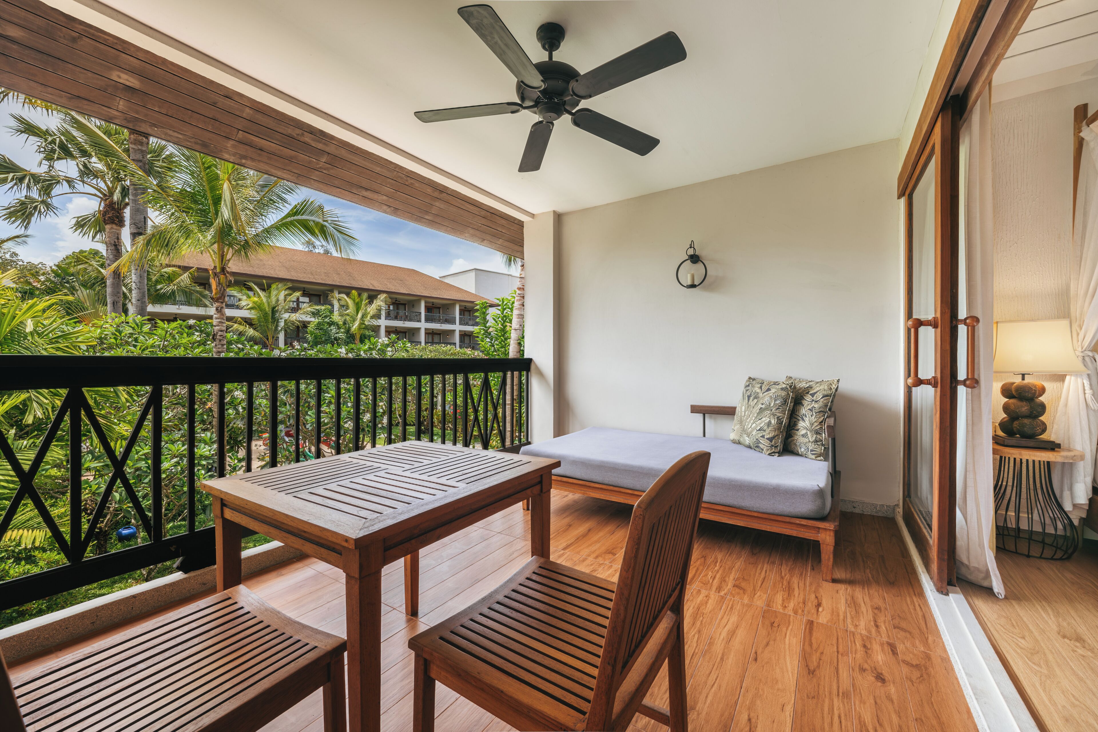 Uma varanda com mesa e cadeiras de madeira, um sofá, um ventilador de teto e vista para um prédio com telhado vermelho.