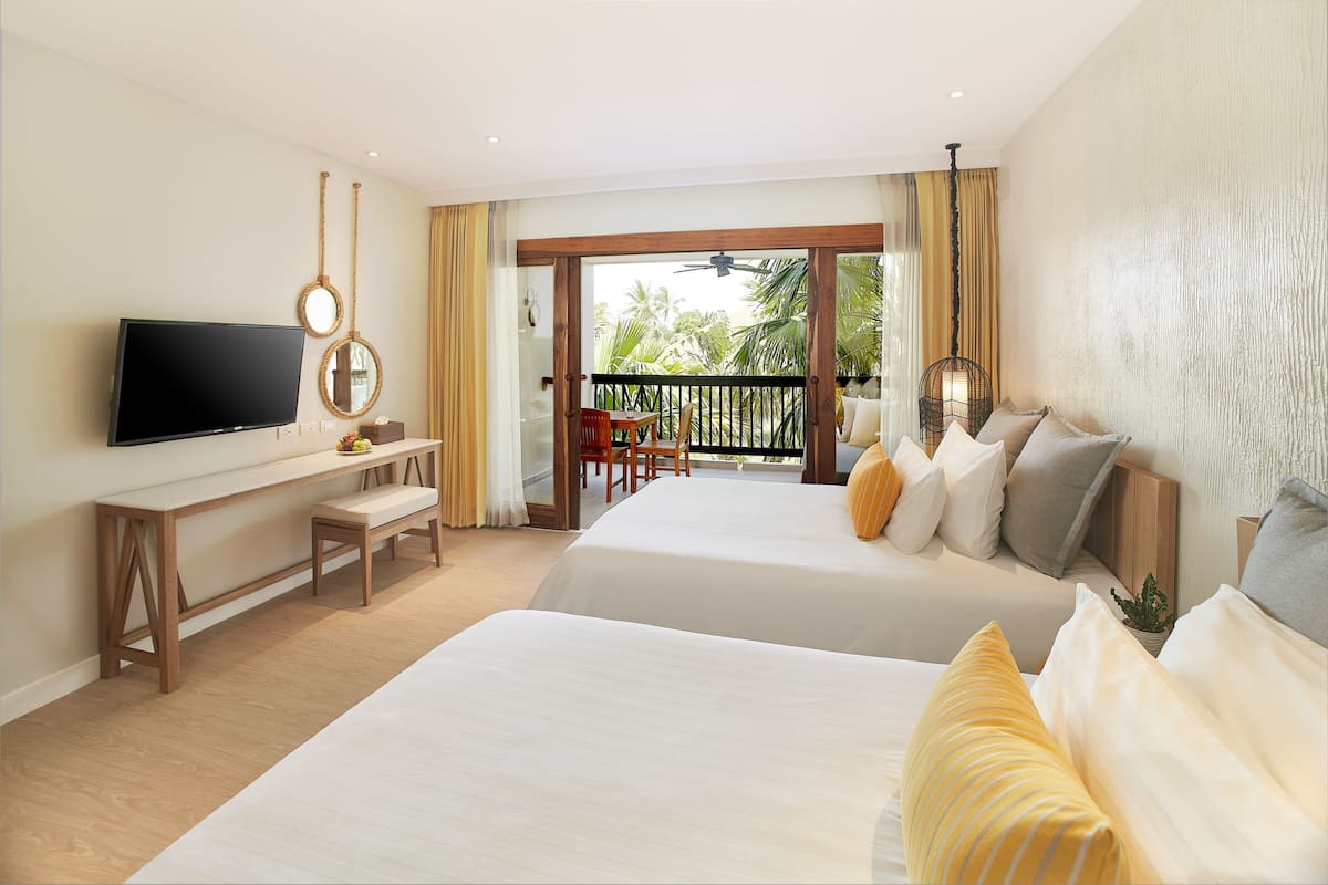 Habitación de hotel con dos camas, televisión, escritorio y balcón con vistas a palmeras.