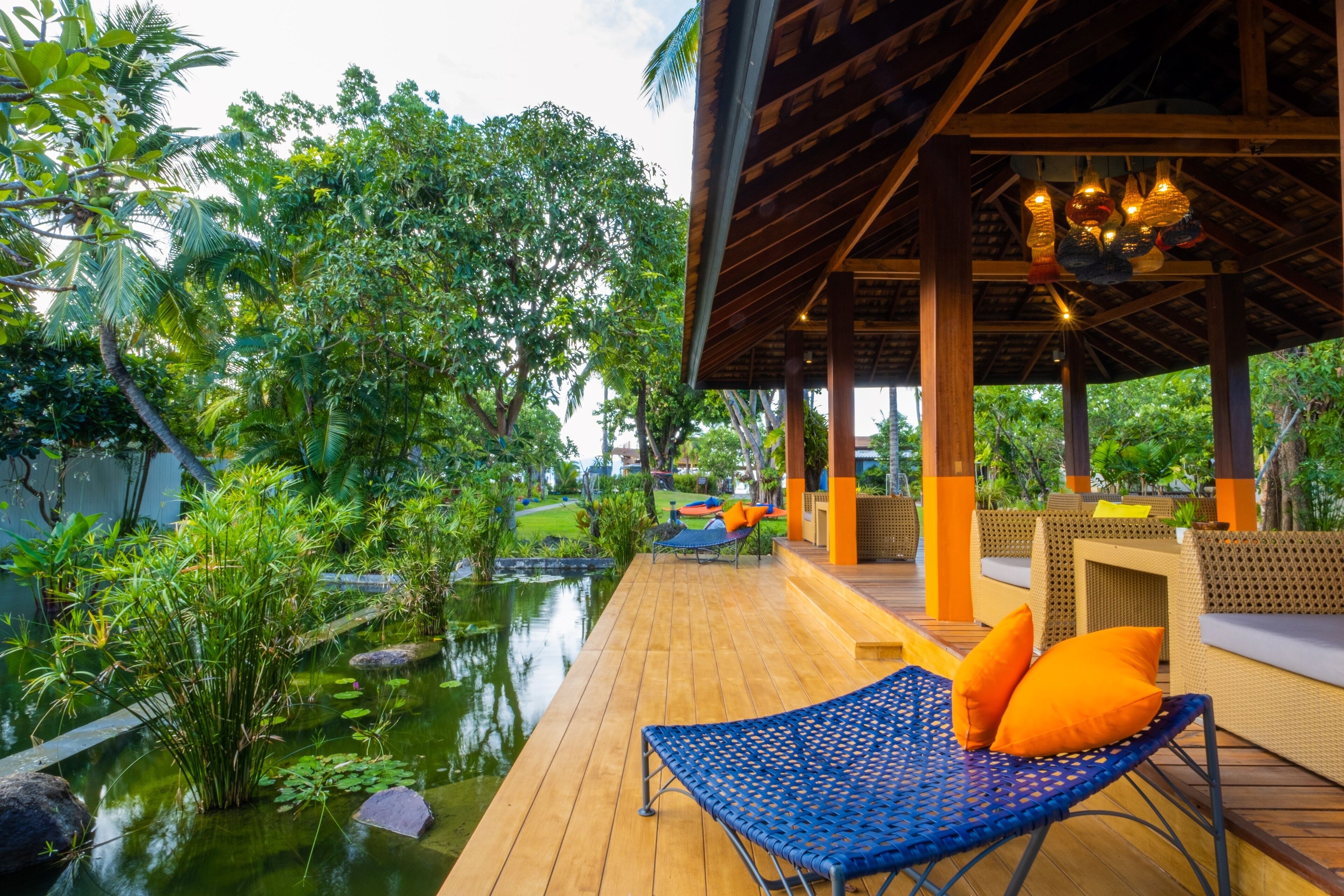 A wooden deck with a lounge chair and cushions, surrounded by lush greenery and a pond.