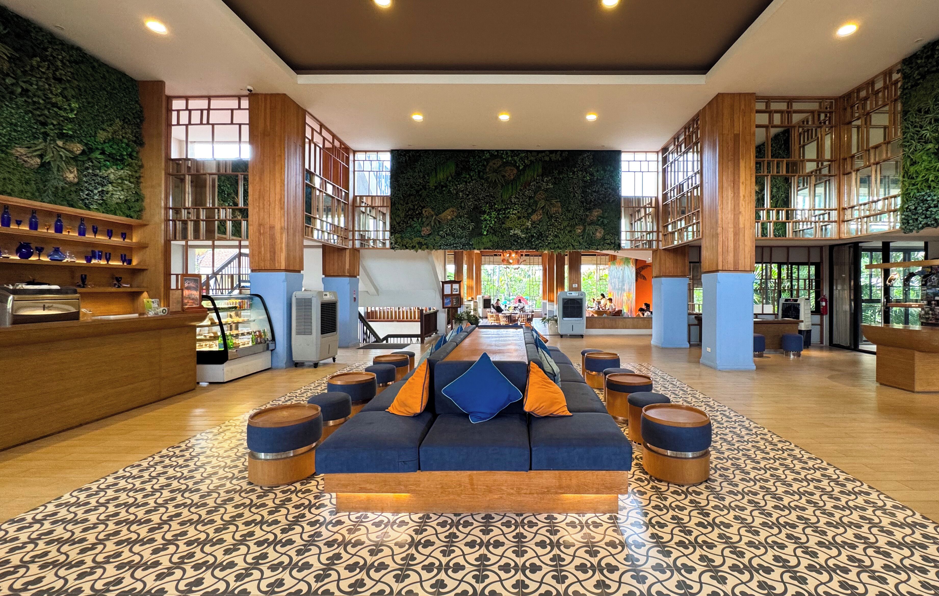 Um hall amplo com um balcão de recepção alongado, uma área de estar central com sofás e pufes, e um chão com padrão.