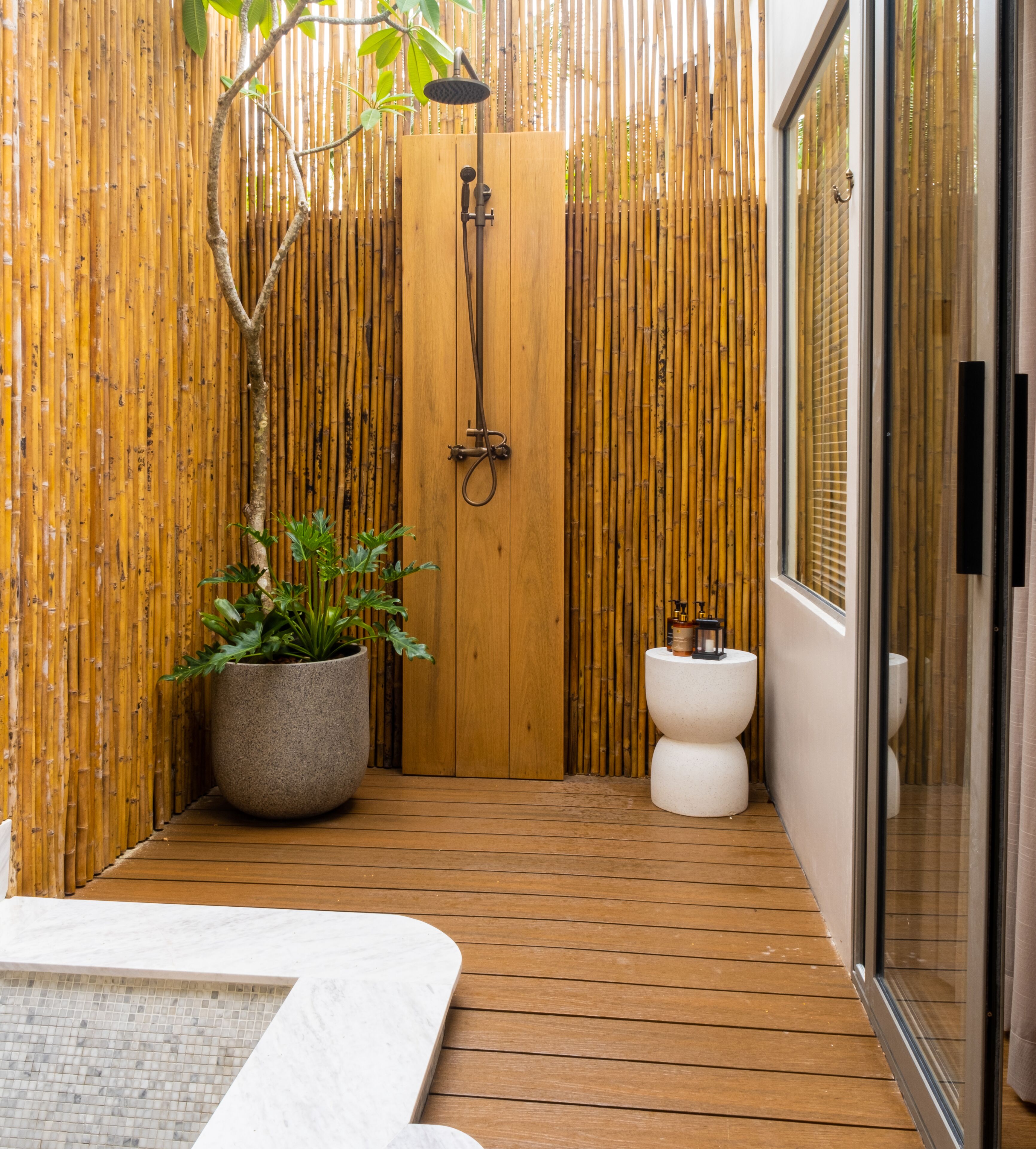Une salle de bain avec un plancher en bois, un mur en bambou, une douche, des toilettes et une plante en pot.