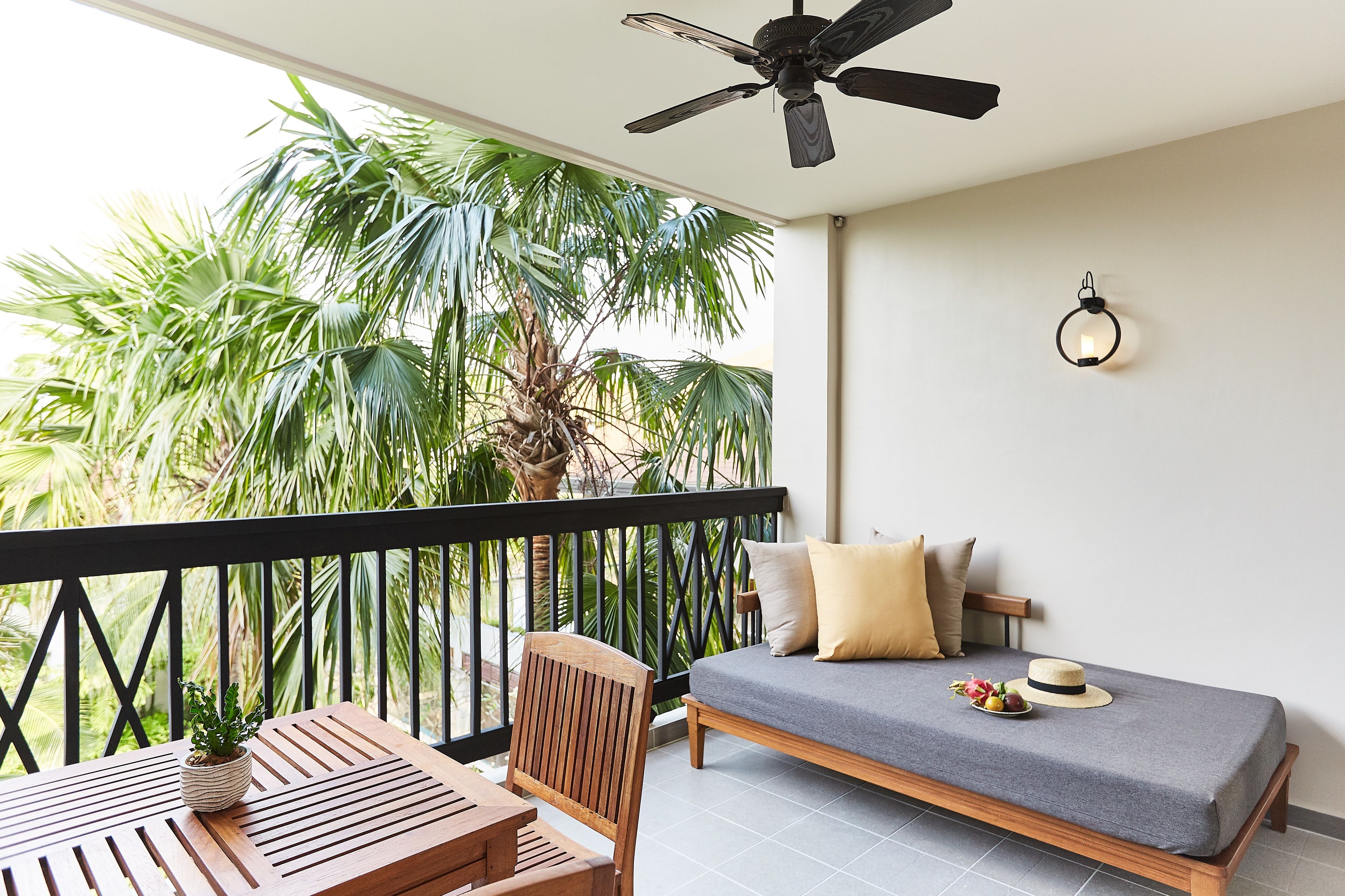 Un balcon avec une table et des chaises en bois, un canapé avec des coussins, un ventilateur de plafond et une vue sur des palmiers.