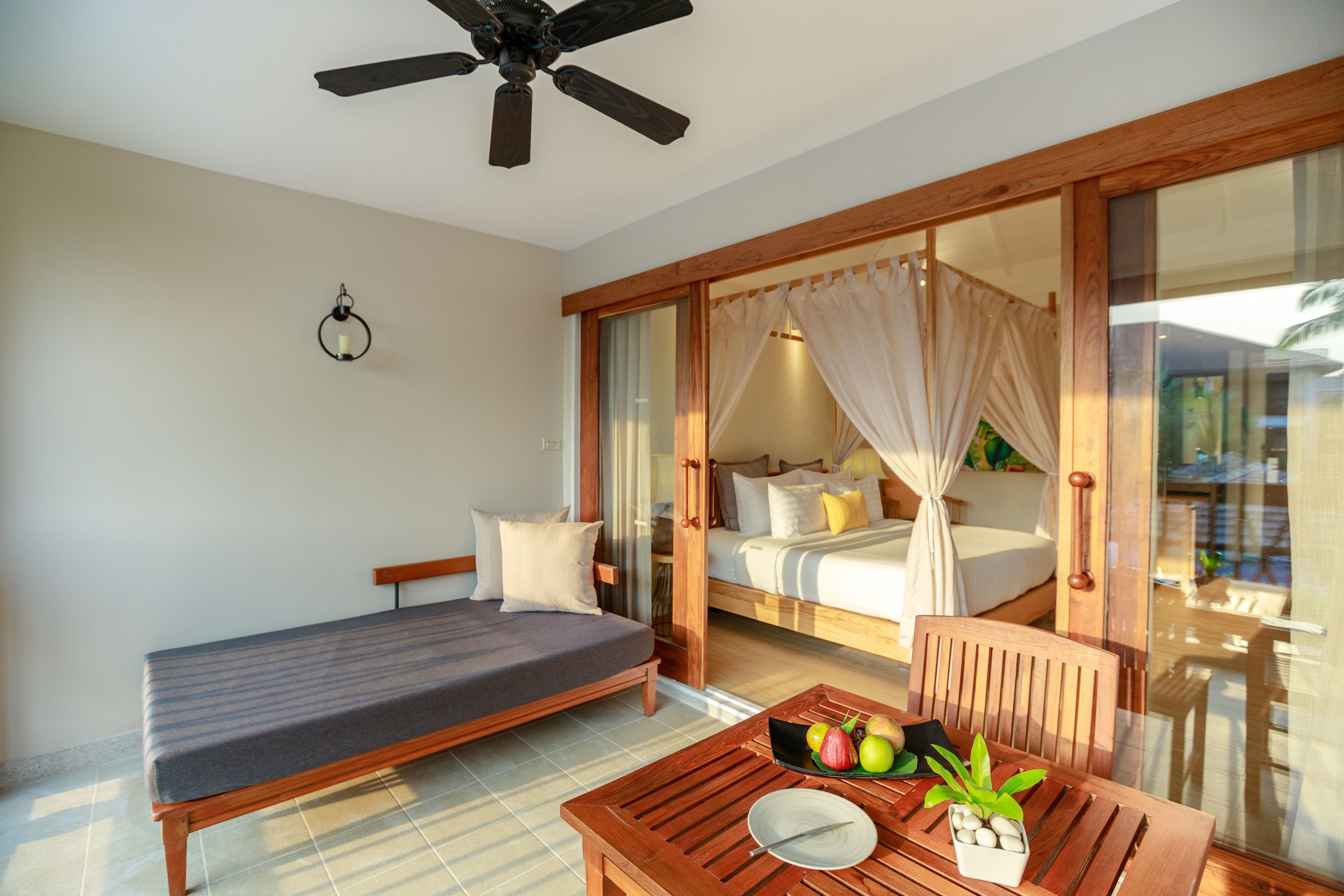 A room with a bed, a sofa, a table with fruits and a plate, and a ceiling fan.