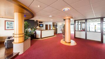 A hotel reception area with a red carpet, wooden columns, a reception desk, and a TV screen.