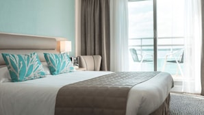 A hotel room with a bed, a nightstand, a lamp, a view of the sea through a sliding door, and a chair by the window. - Hôtel de l’Océan Concarneau (Concarneau)