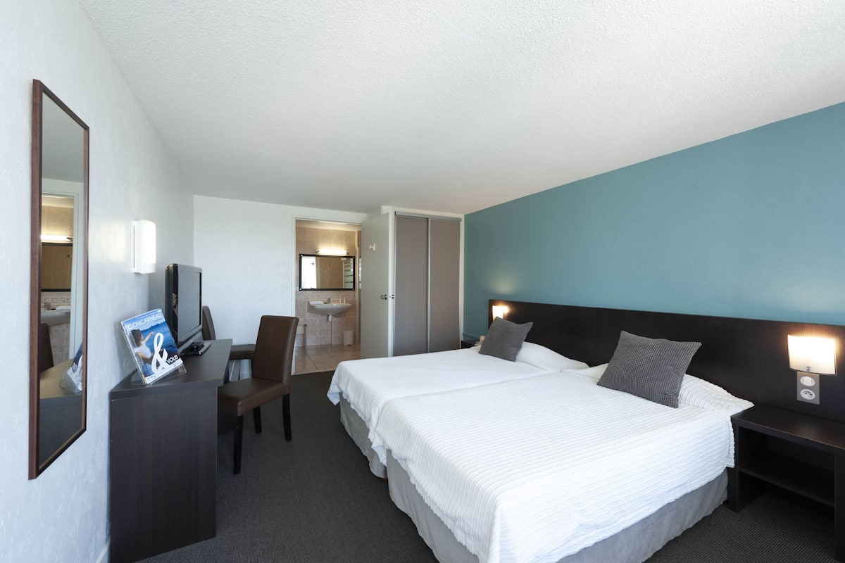 Une chambre d’hôtel avec un grand lit, un bureau avec une chaise, un miroir et une salle de bain visible à travers une porte ouverte.