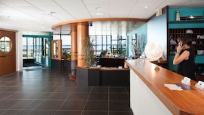 A hotel reception area with a curved wooden counter, a receptionist, and a view of the sea through large windows. - Hôtel de l’Océan Concarneau (Concarneau)