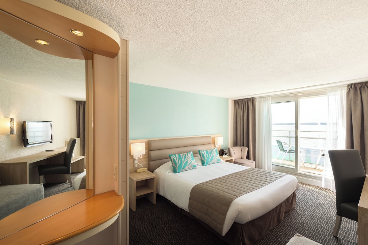 Une chambre d’hôtel avec un grand lit, un bureau, une chaise, une télévision et une vue sur la mer par la fenêtre.