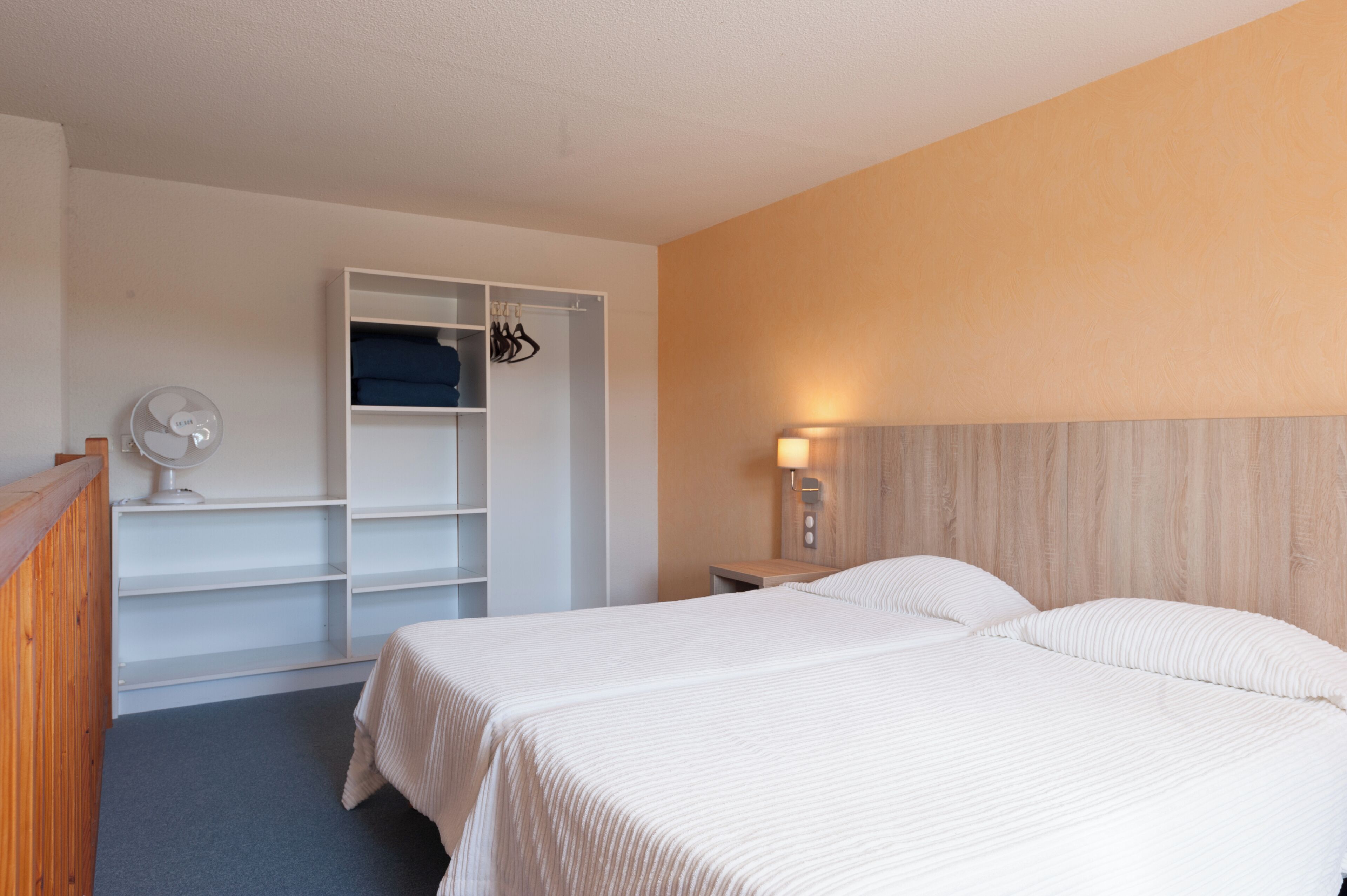 A hotel room with two beds, a wardrobe, a bedside table with a lamp, and a ceiling fan.