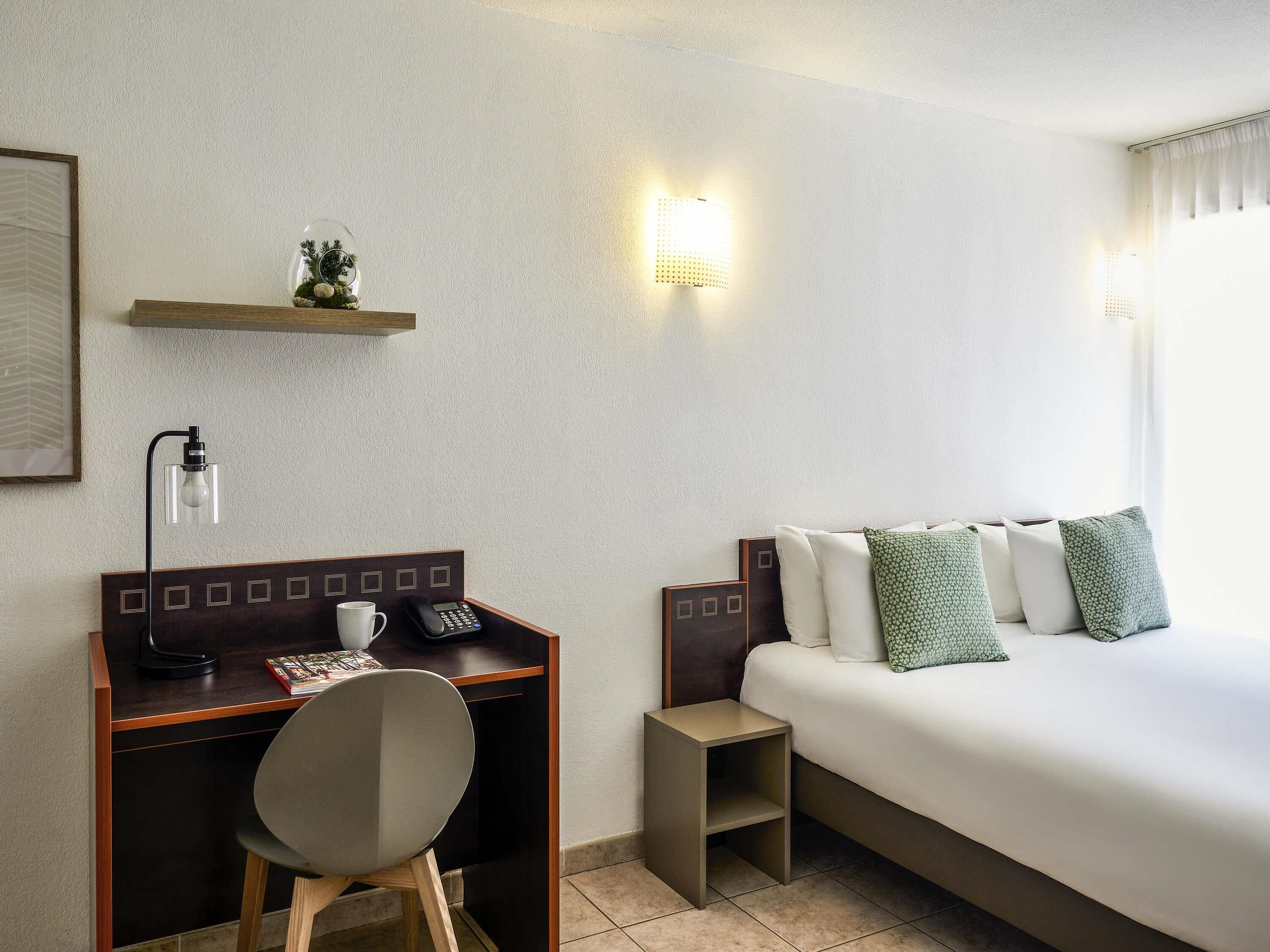 Une chambre d’hôtel comprenant un lit, un bureau avec une chaise, un téléphone, une lampe et une petite étagère avec des objets décoratifs.