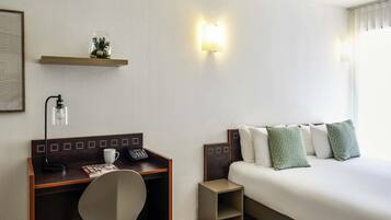 Una habitación de hotel con cama, escritorio con silla, teléfono, lámpara y un estante con adornos.