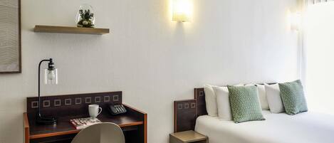 A hotel room with a bed, a desk with a chair, a telephone, a lamp, and a small shelf with decorative items.