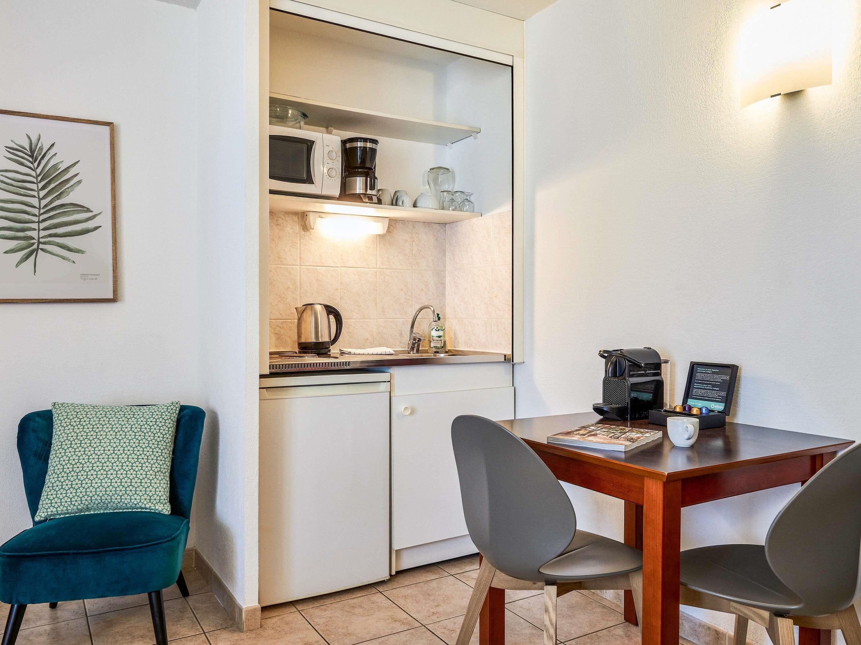 Een compacte keuken met een magnetron, koffiezetapparaat en een vaatwasser. Er is een eettafel met twee stoelen en een teal-kleurige stoel.