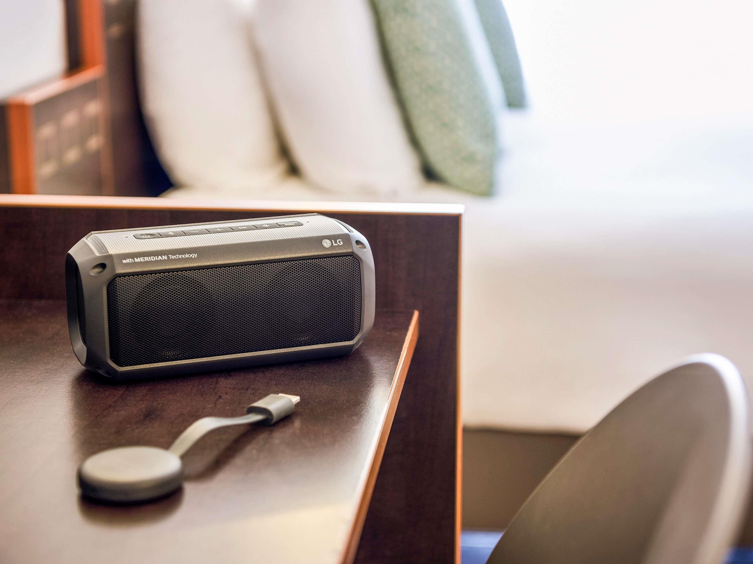 A portable speaker with a charging cable on a wooden table in a hotel room.